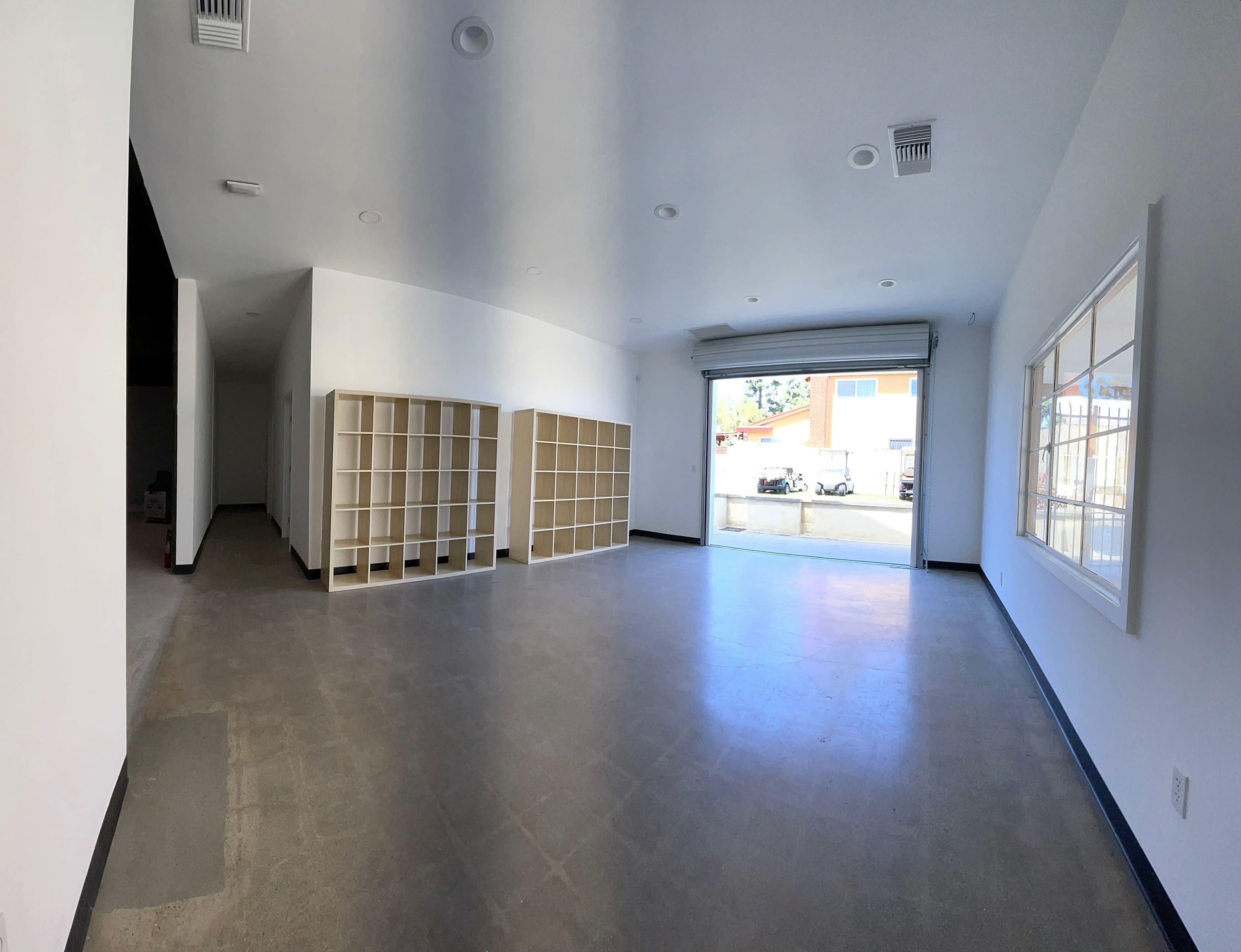 A spacious, empty interior with white walls, concrete flooring, and several shelving units along one side, with a wide open garage door leading outside.