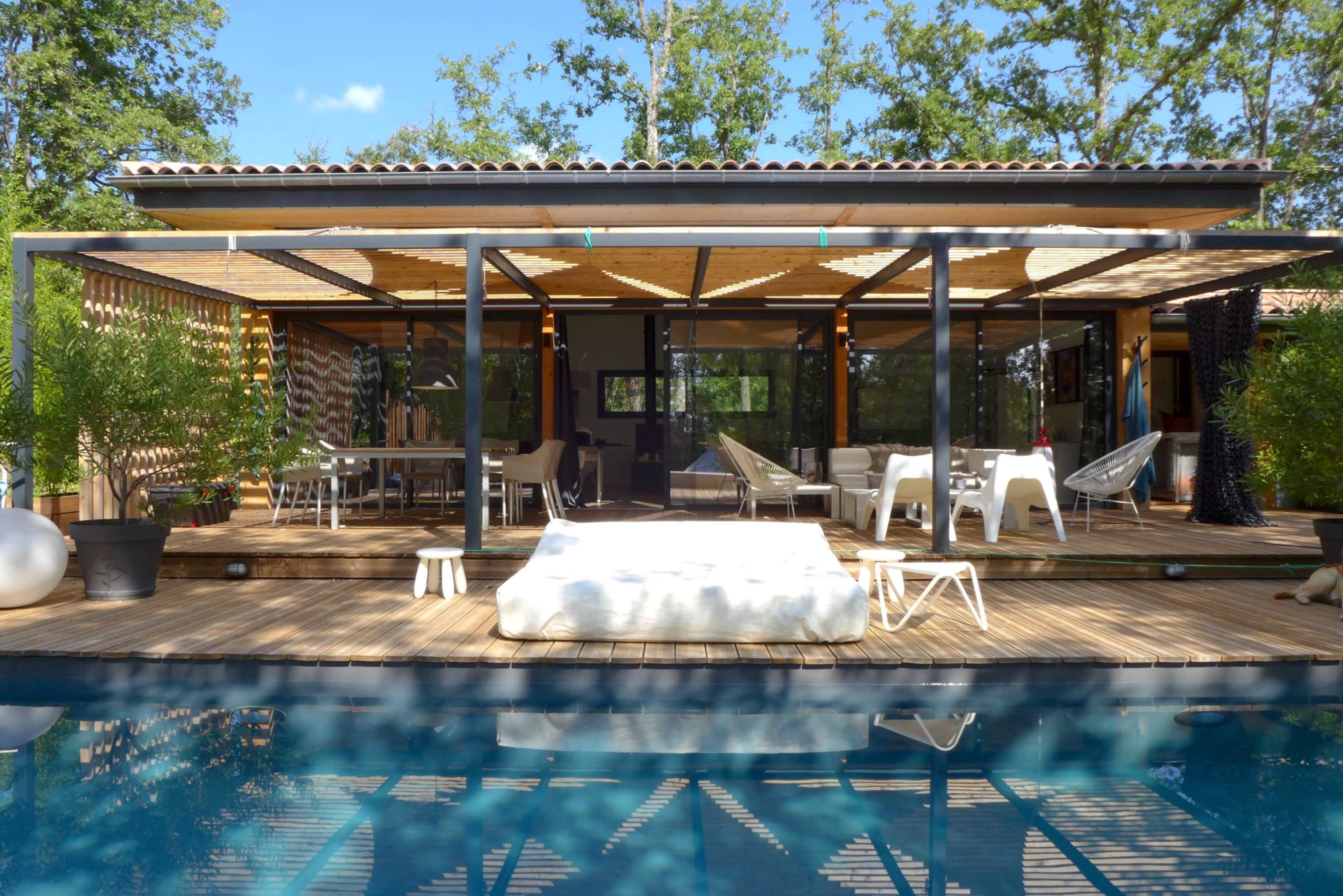 A modern house with a wooden deck and glass walls overlooks a swimming pool surrounded by trees.