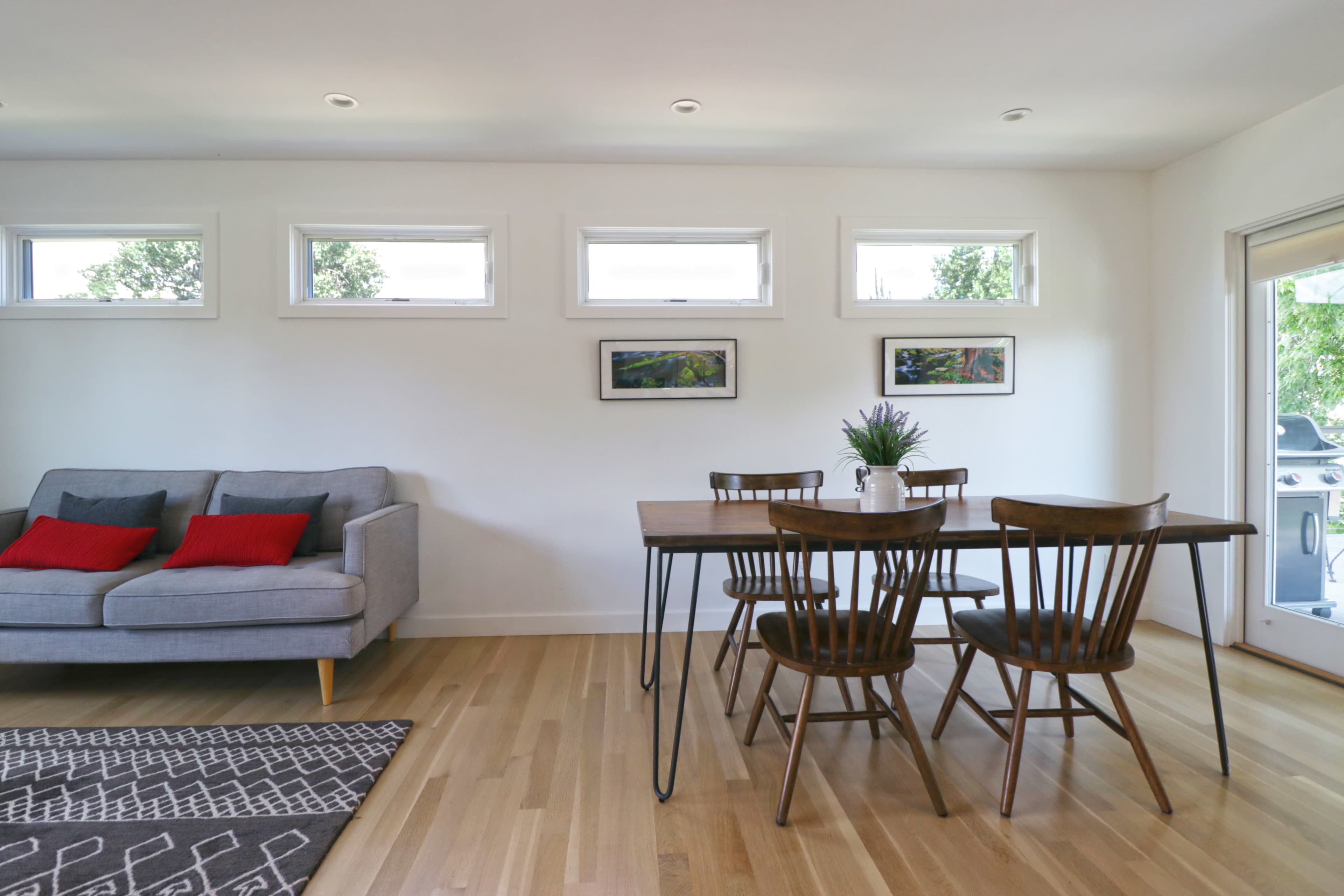 A modern dining area with a wooden table surrounded by chairs, a gray sofa, and large windows allowing natural light.