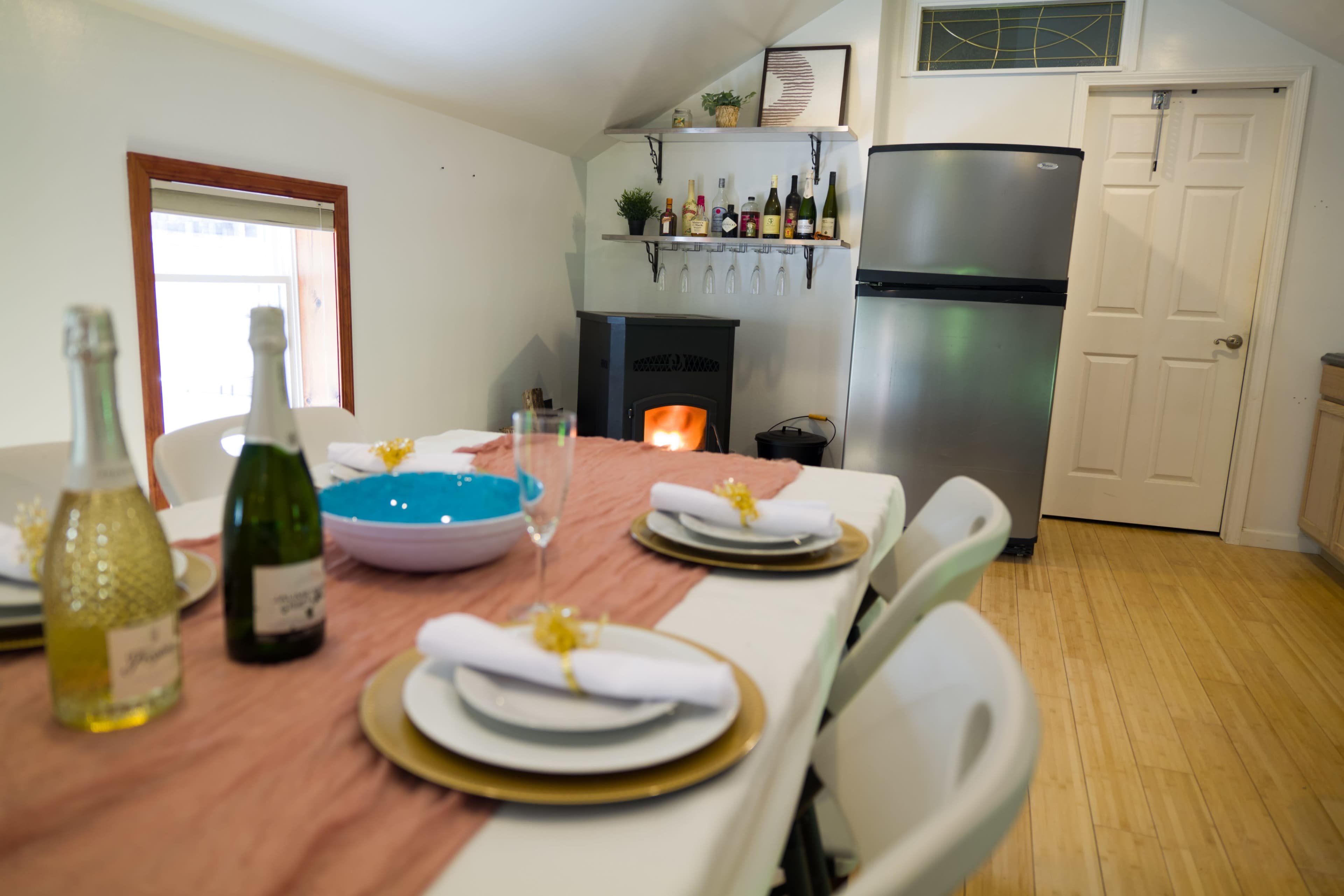 A dining table set with white napkins and decorative items is positioned in a well-lit kitchen featuring a refrigerator and a small stove.