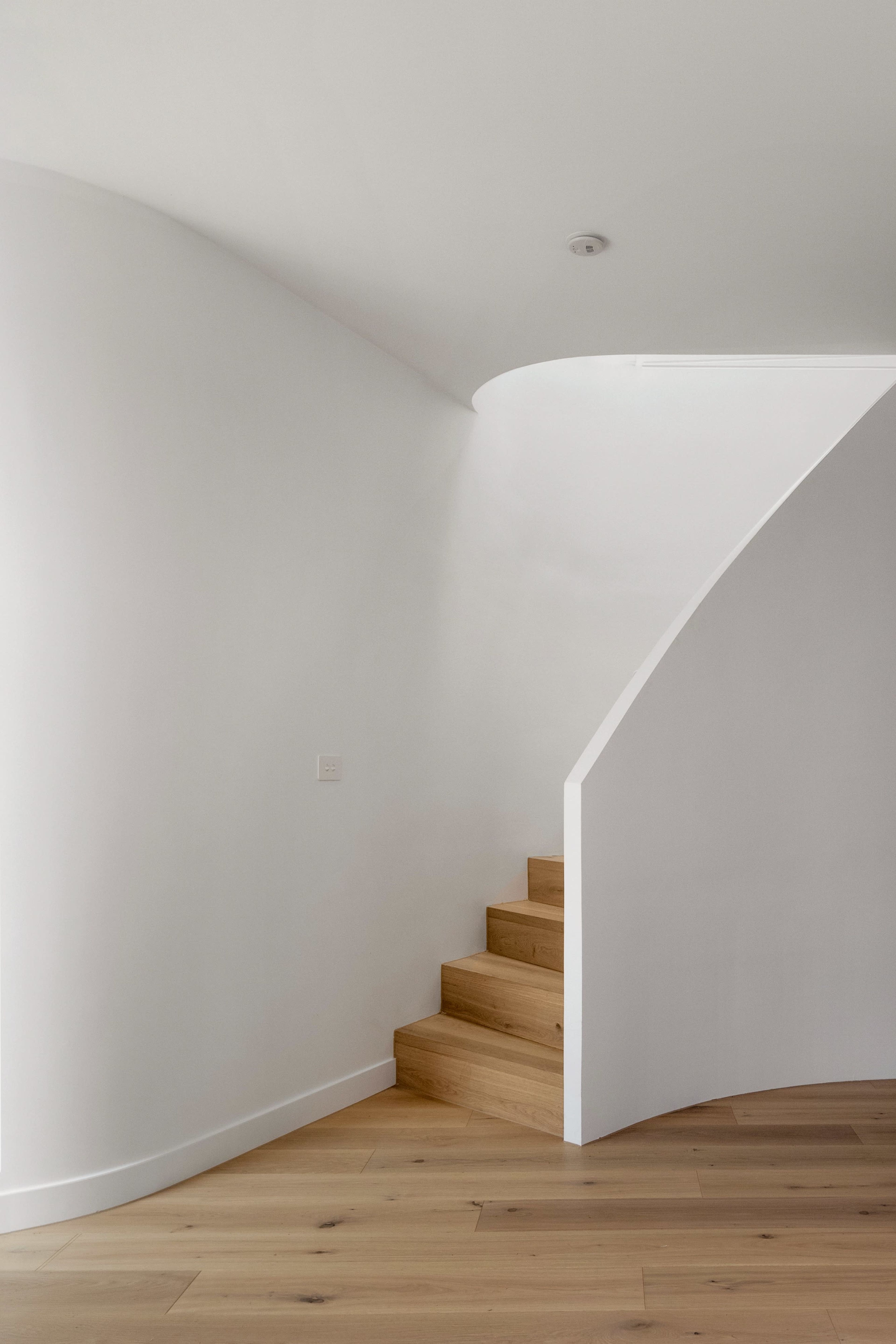 A curved wall with a staircase made of wooden steps leads upward in a minimalist, white interior space.