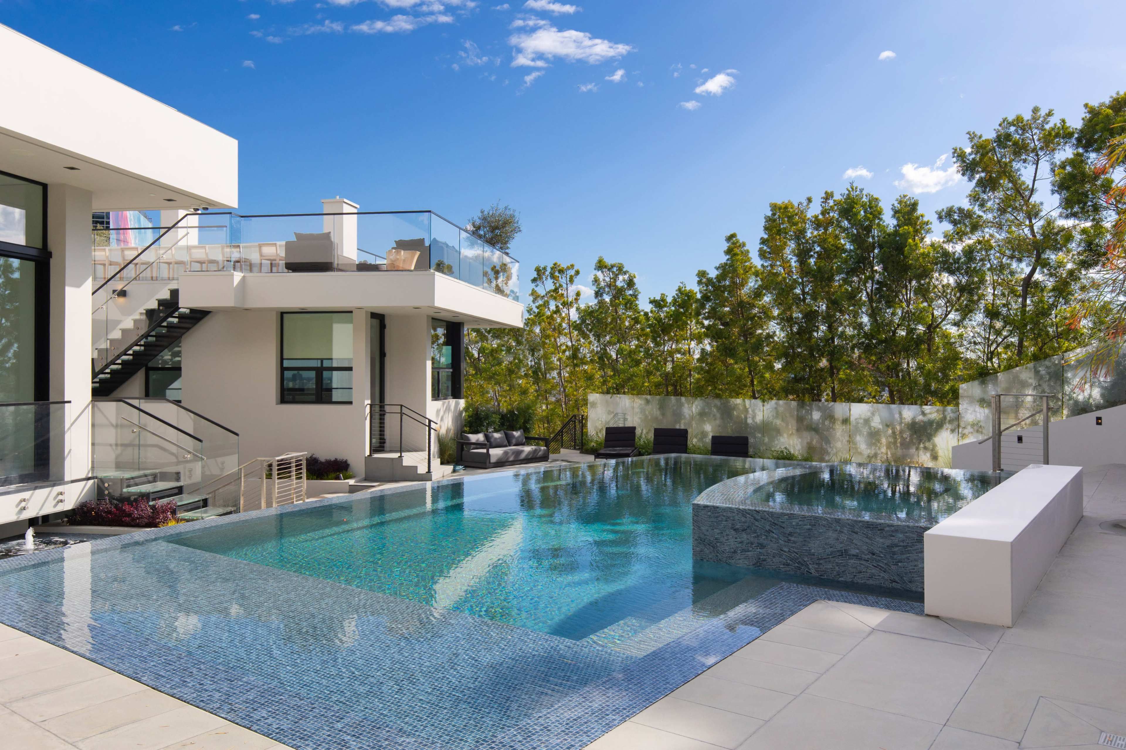 The image shows a modern home featuring a large swimming pool with an infinity edge, surrounded by greenery and outdoor seating areas.