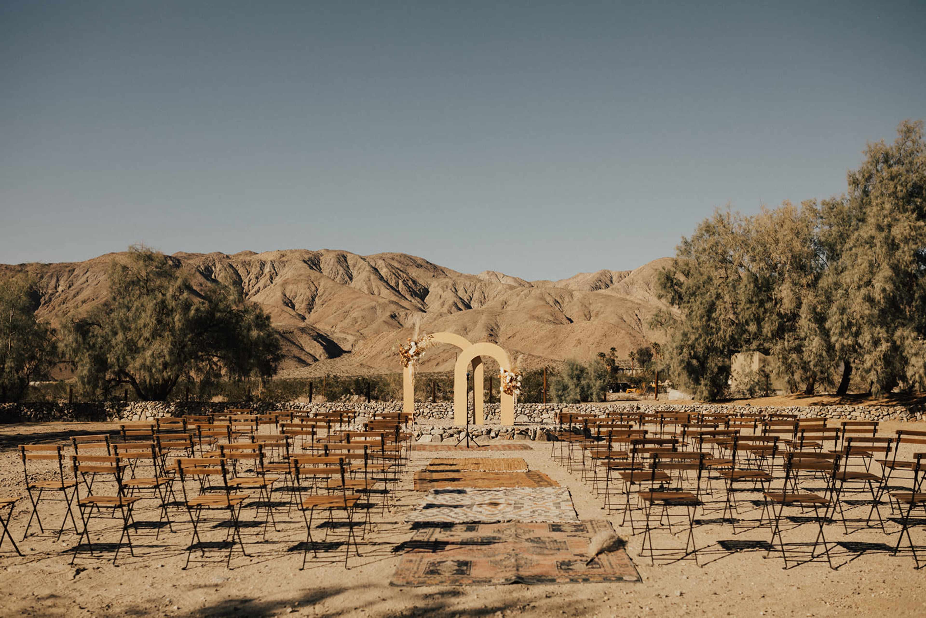 A wedding ceremony setup with rows of wooden chairs facing a decorated arch in a mountainous desert landscape.