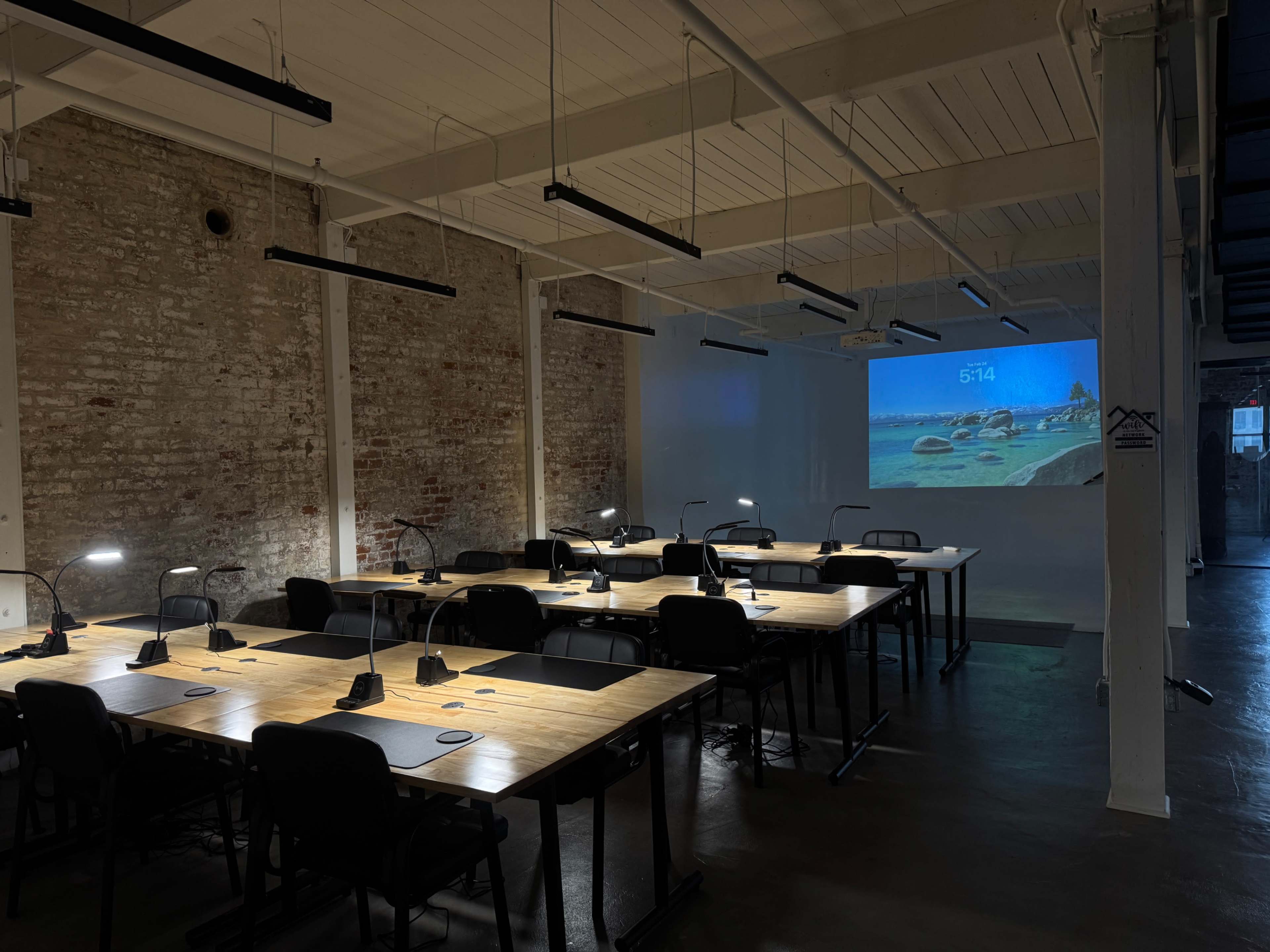 The image shows a modern, spacious office with multiple workstations illuminated by task lamps and a projector displaying a scenic image on a white wall.