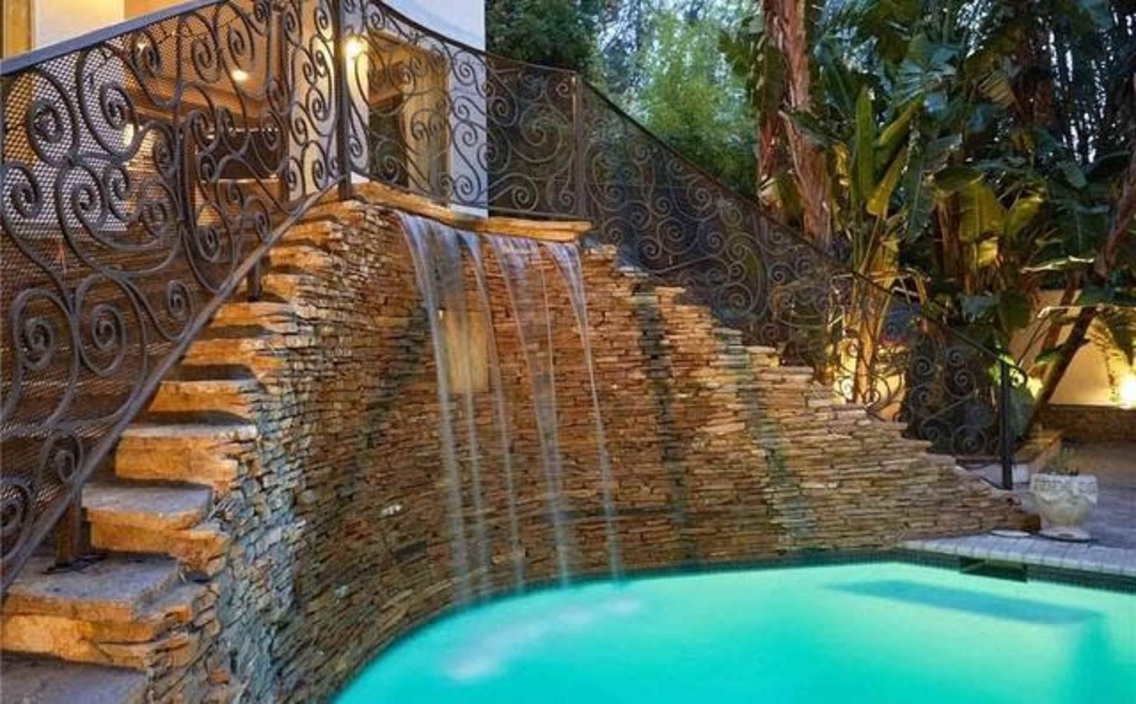 A waterfall cascades down stone steps into a turquoise pool surrounded by lush greenery and decorative metalwork.