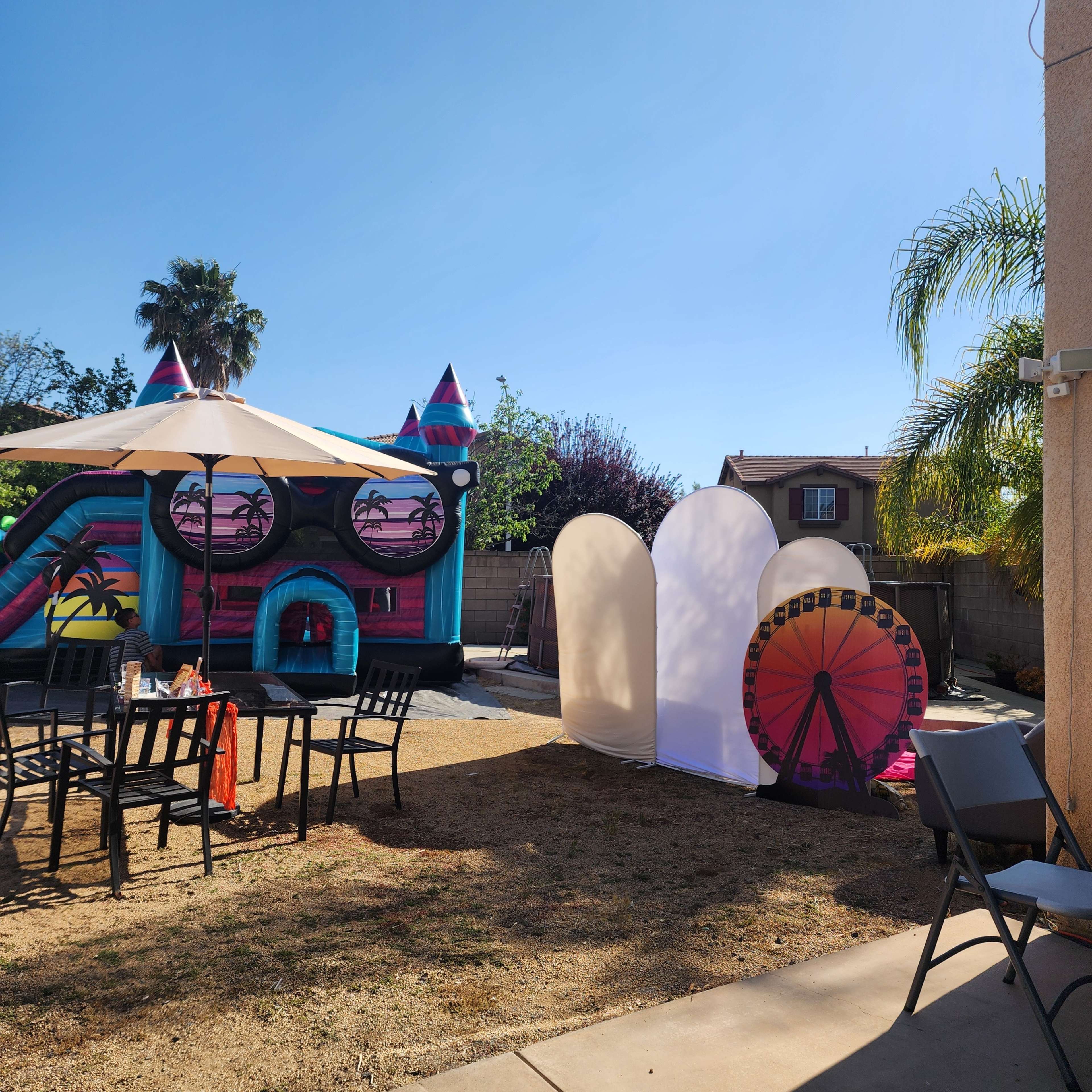 A colorful inflatable bounce house featuring sunglasses and palm trees is set up in a backyard with a seating area and a large white backdrop nearby.