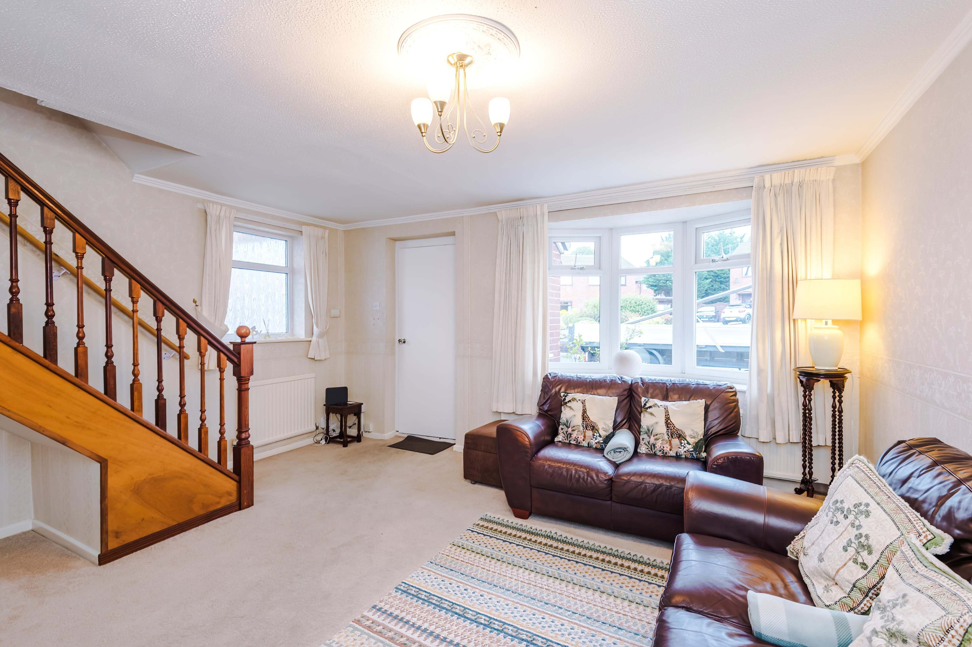 The image shows a living room with a staircase, a brown leather sofa, a patterned rug, and two large windows letting in natural light.