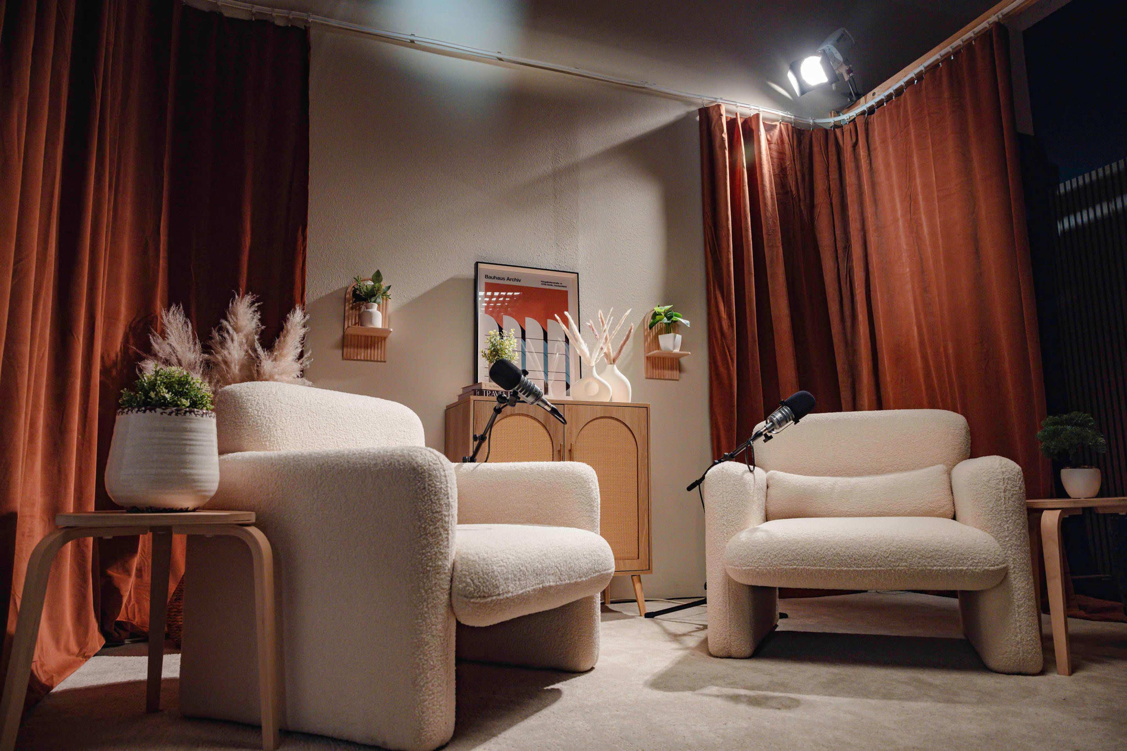 A cozy, minimalist interview setup with two white armchairs, microphones on stands, and decorative plants against a backdrop of orange curtains.