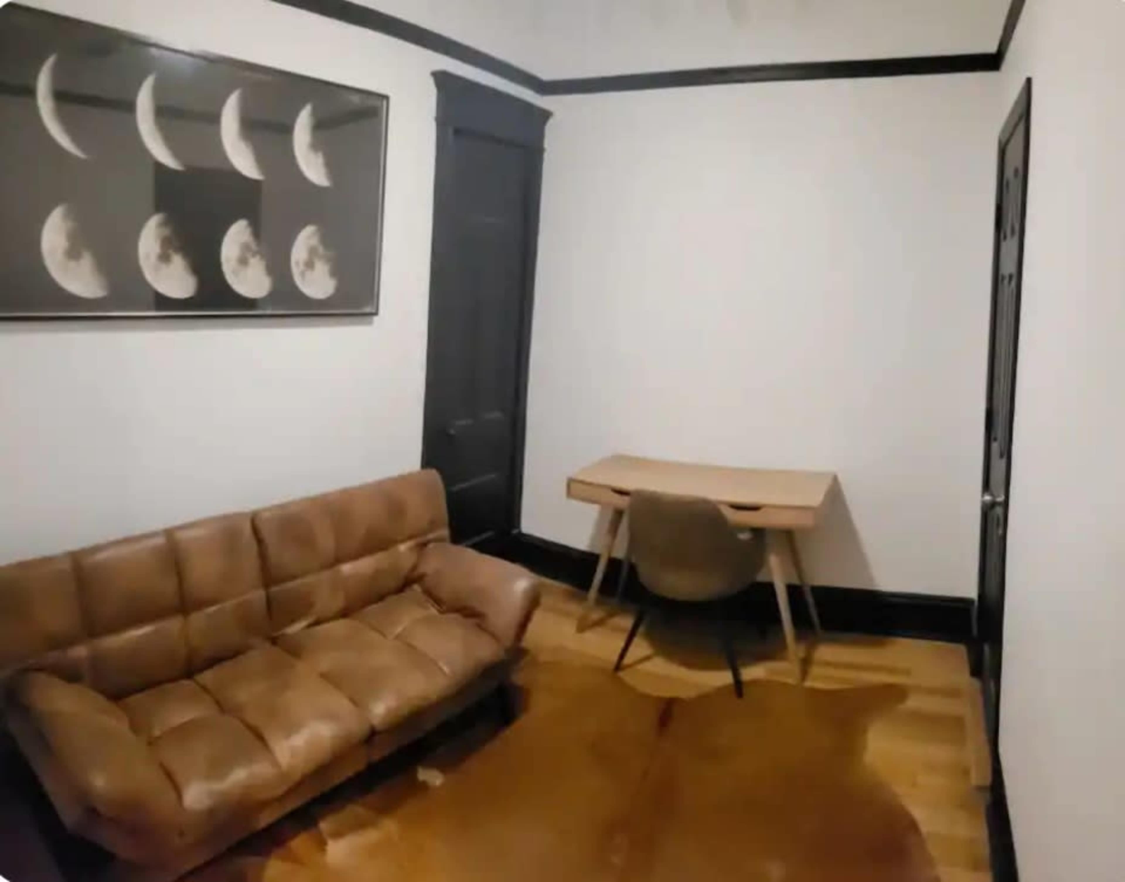 The image shows a sparse room with a brown leather sofa, a wooden desk, and a framed artwork of moon phases on the wall.