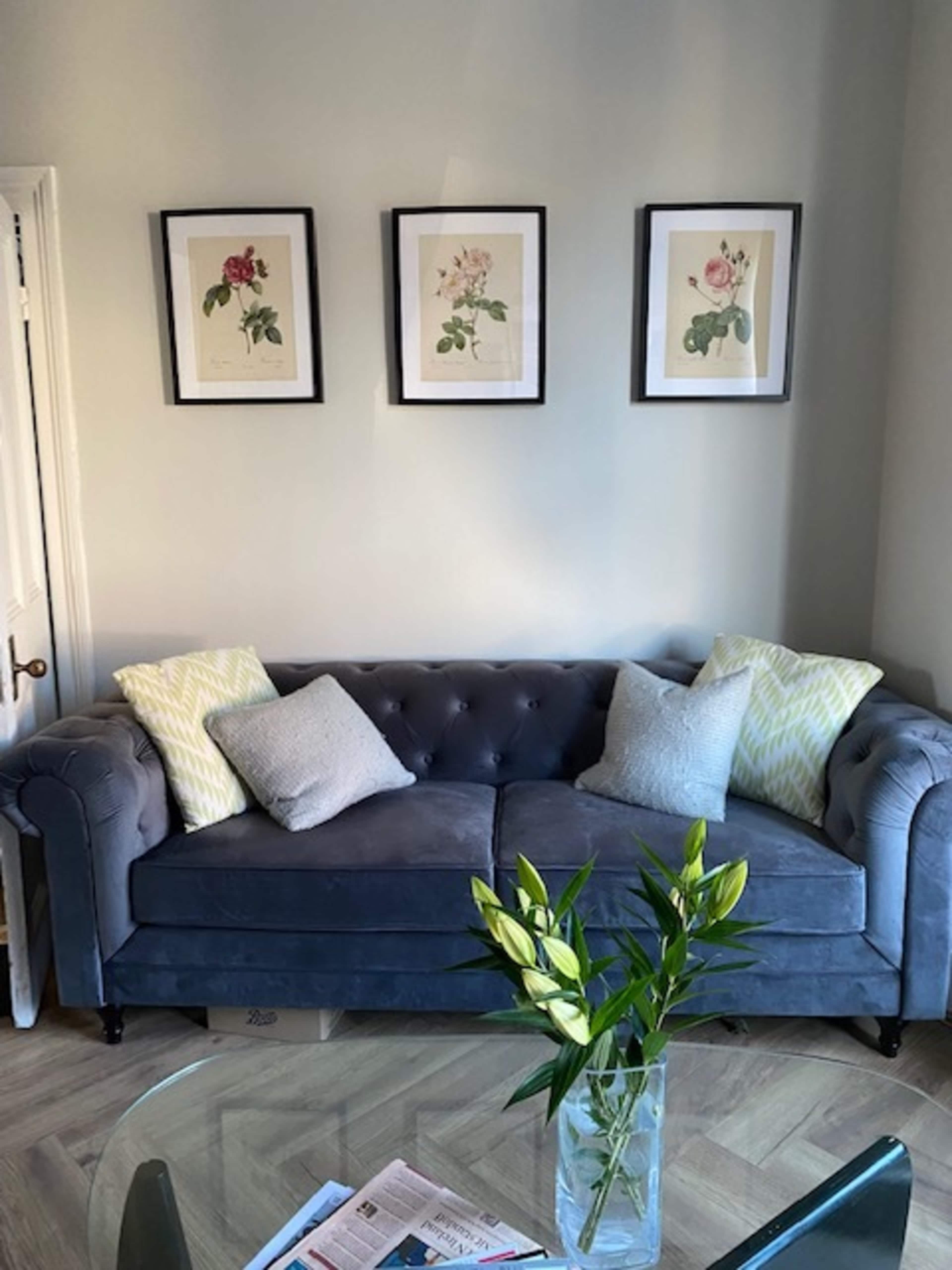 A gray tufted sofa with patterned pillows is positioned against a light-colored wall, adorned with three framed botanical illustrations above it.