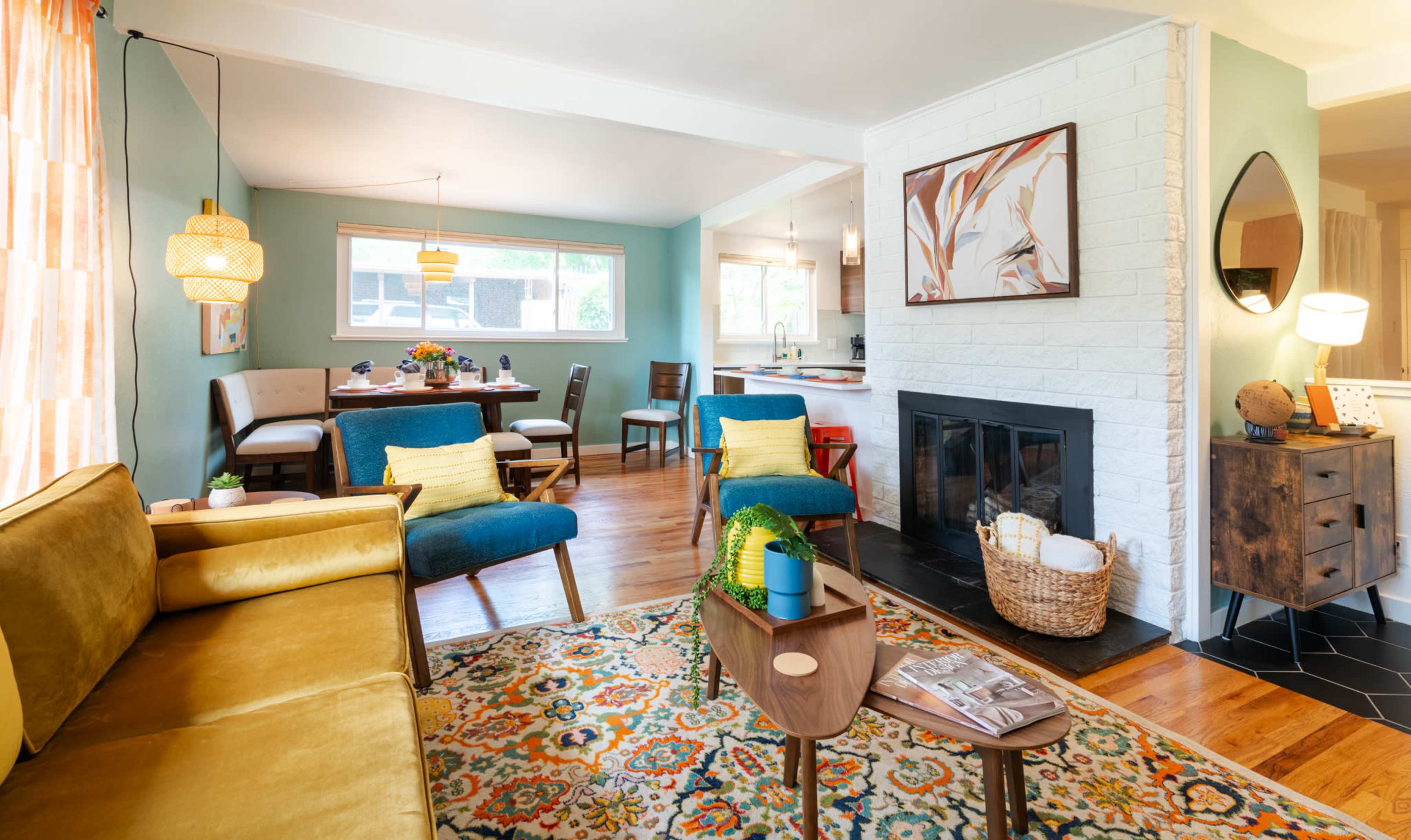 A modern living room features a yellow sofa, blue accent chairs, a patterned rug, and a fireplace, with a dining area visible in the background.
