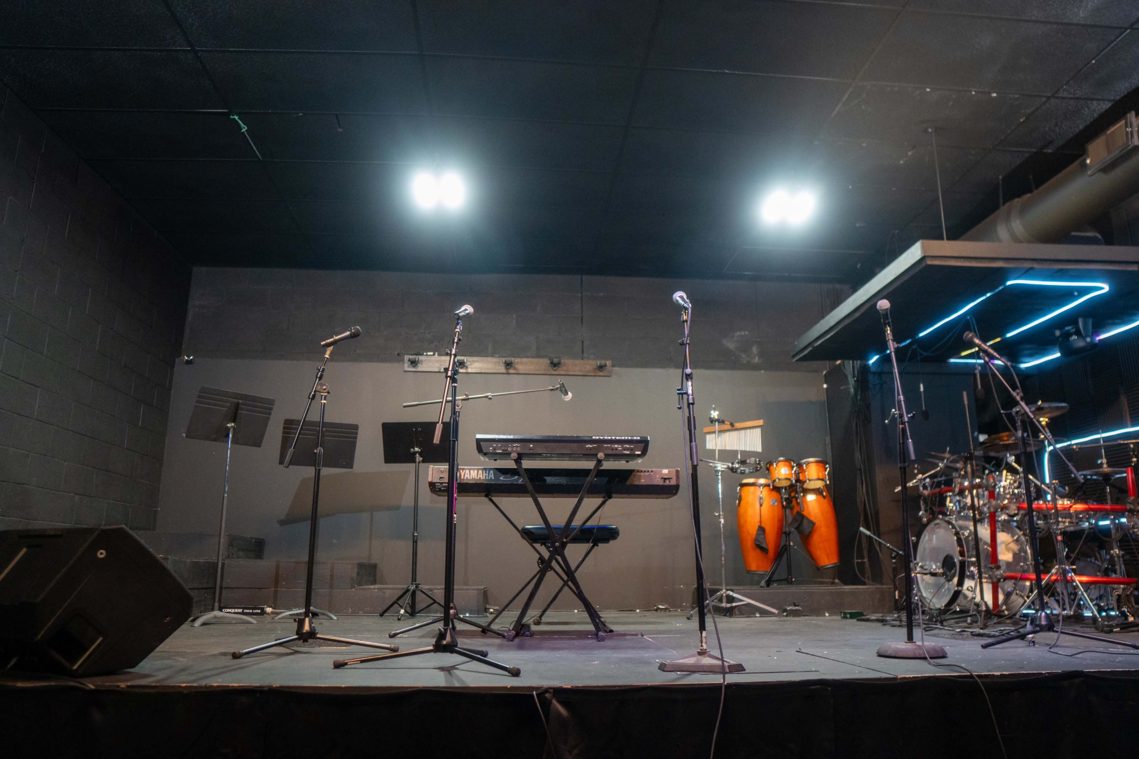 The image shows a stage set up with a keyboard, several microphones, and various percussion instruments, including congas and a drum kit, against a dark backdrop.
