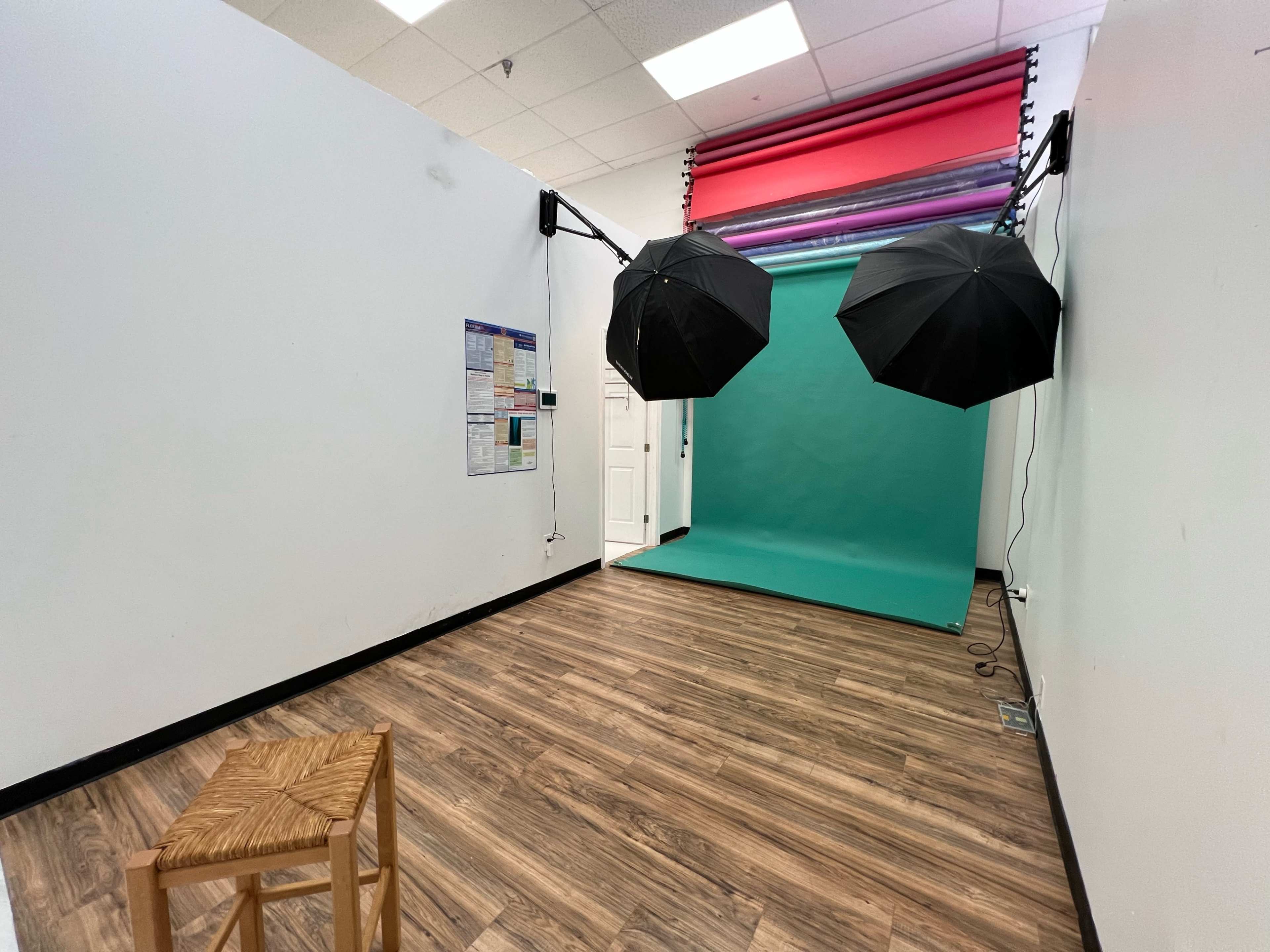 A small photography studio with a teal backdrop, two black umbrellas for lighting, and a wooden stool.