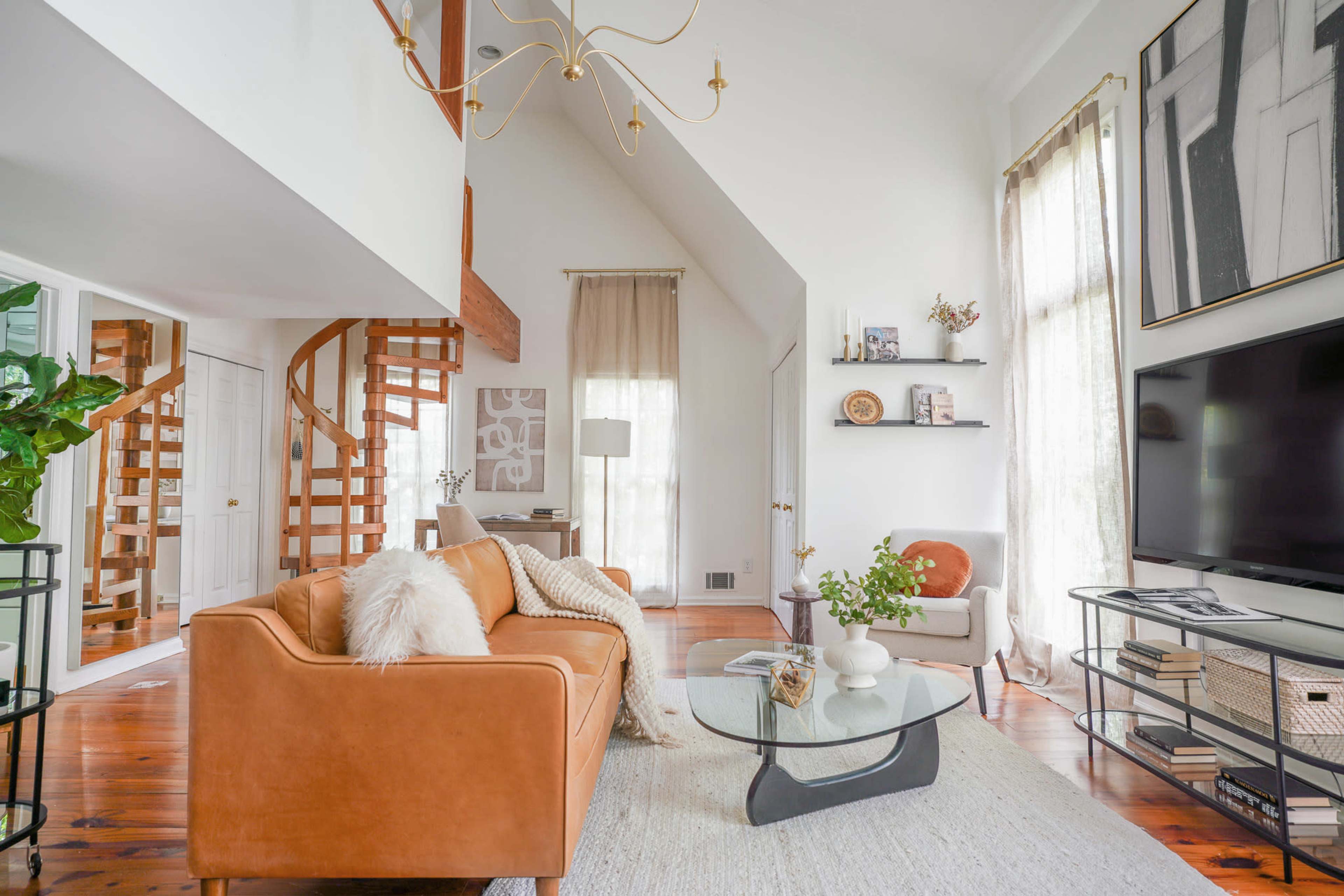 A modern living room with a spiral staircase, a brown leather sofa, a glass coffee table, and large windows allowing natural light to enter.
