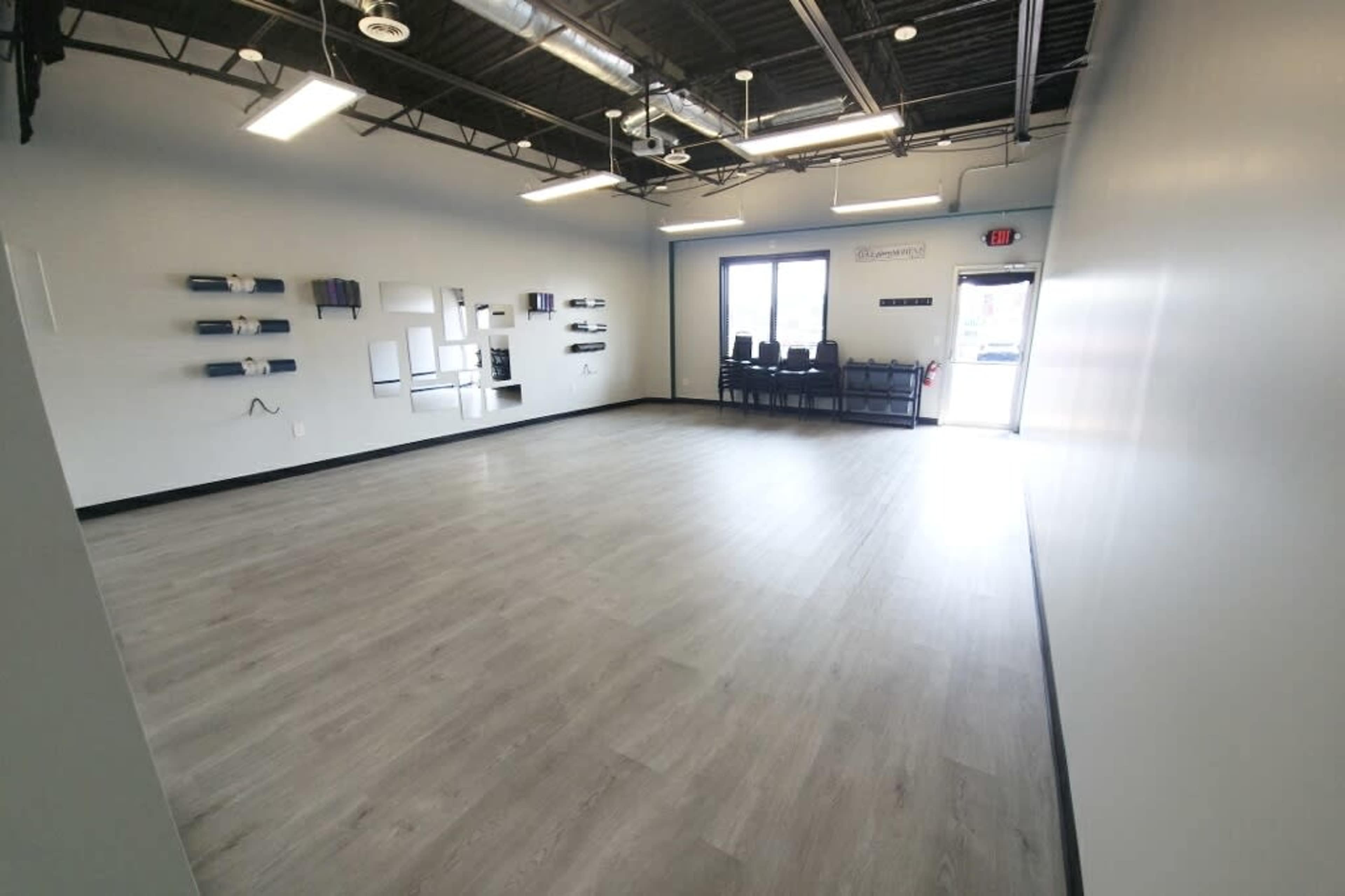 An empty, well-lit room with gray flooring, white walls, and a large window showing rows of chairs stacked near the entrance.