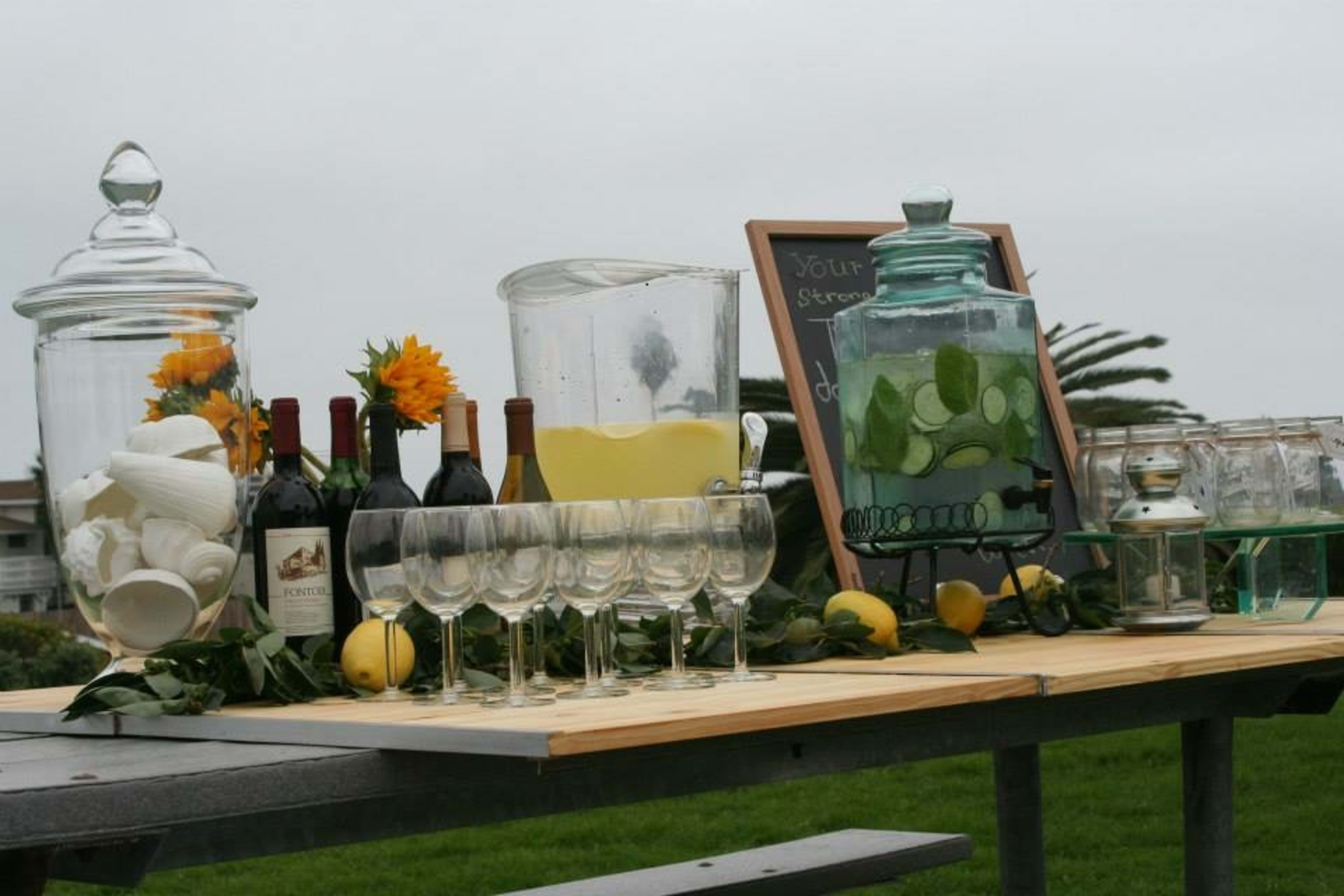 A table is set up outdoors with glass jars, bottles of wine, and glasses, accompanied by lemons and greenery.