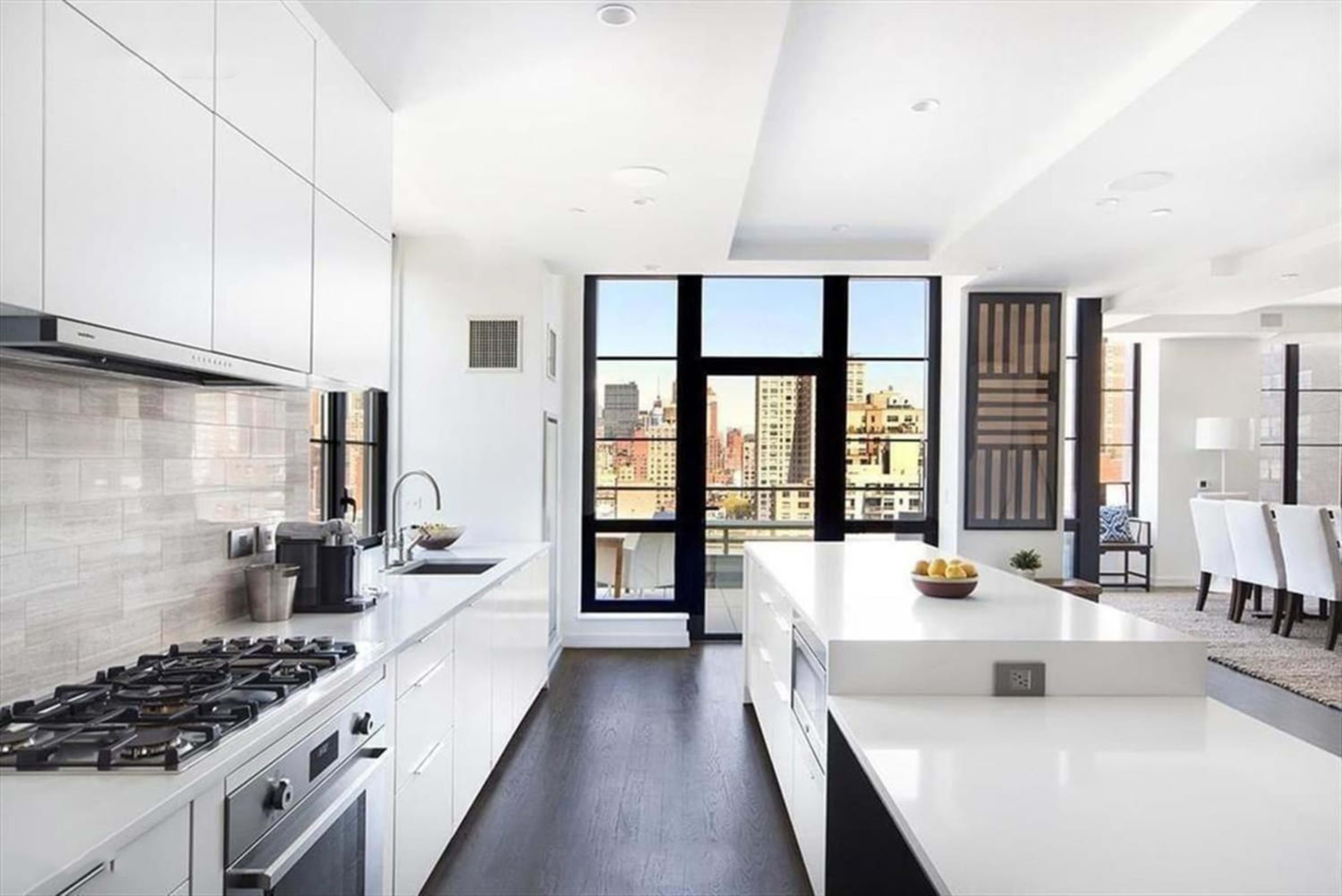 A modern kitchen features sleek white cabinets, a large island with a sink, and floor-to-ceiling windows overlooking a cityscape.