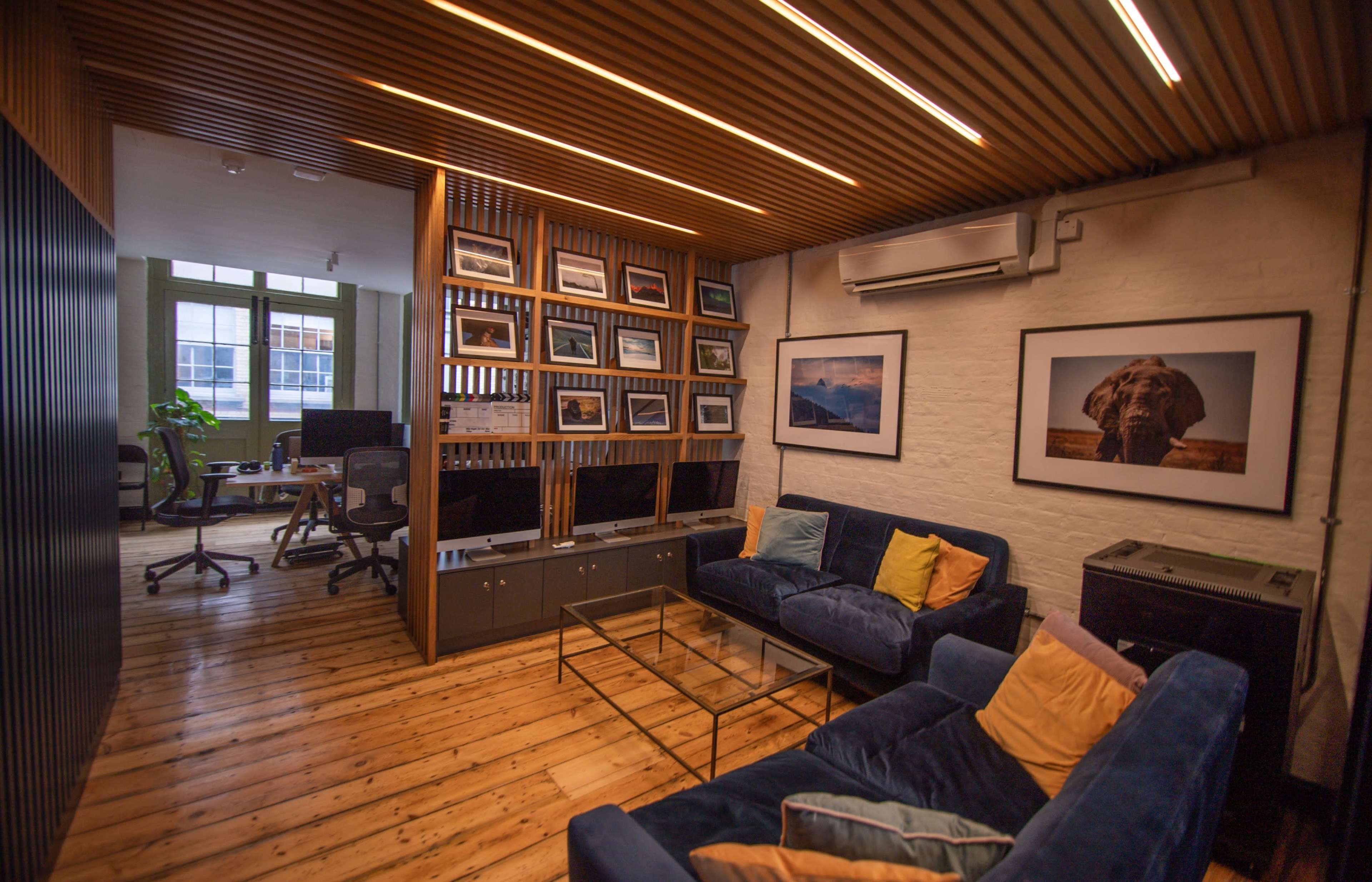 A modern office space with wooden flooring, featuring a lounge area with plush sofas and a wall of framed photographs alongside workstations in the background.