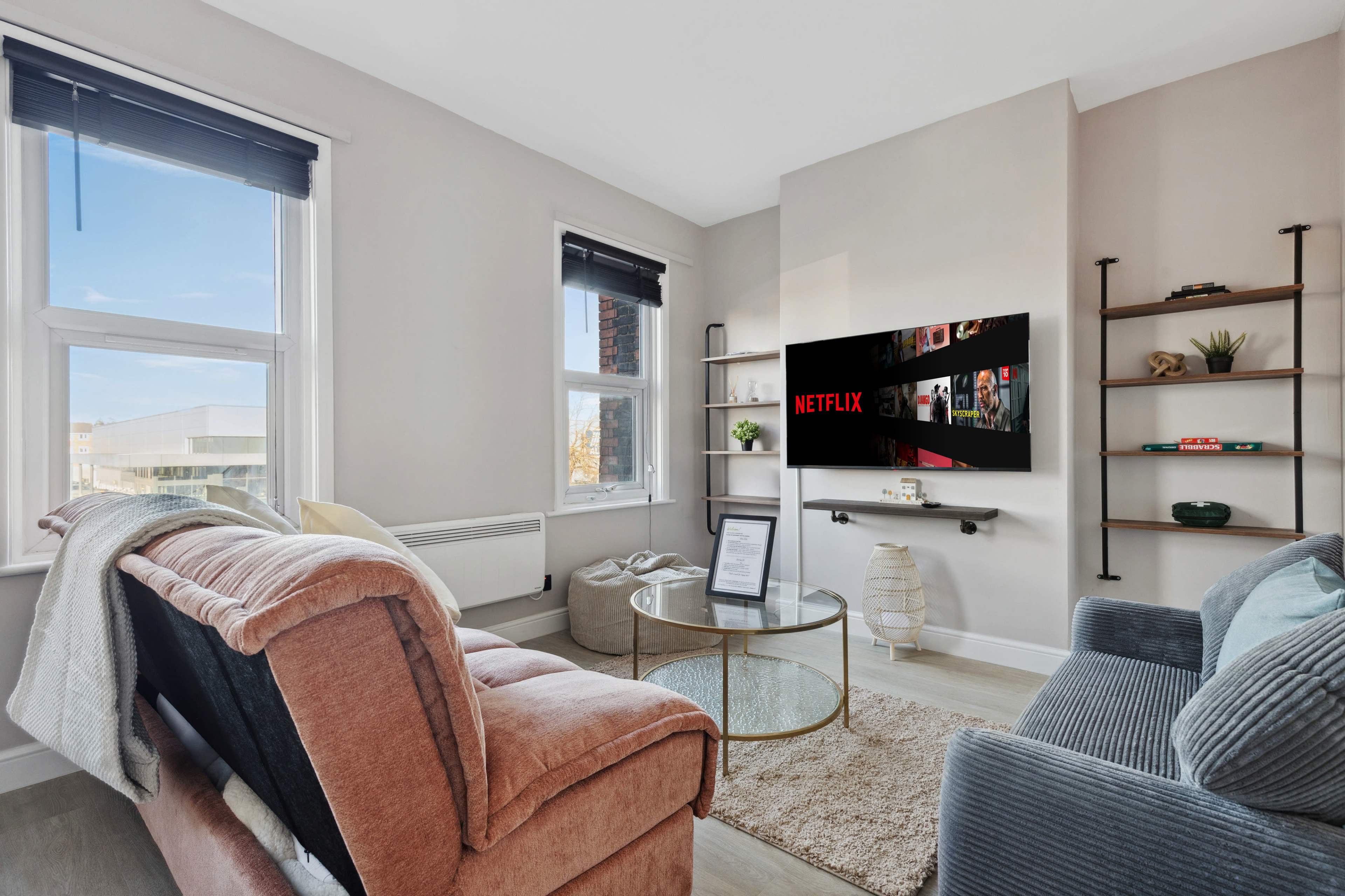 The living room features a modern layout with a pink sofa and a gray armchair, a round glass coffee table, and a wall-mounted TV displaying the Netflix logo.