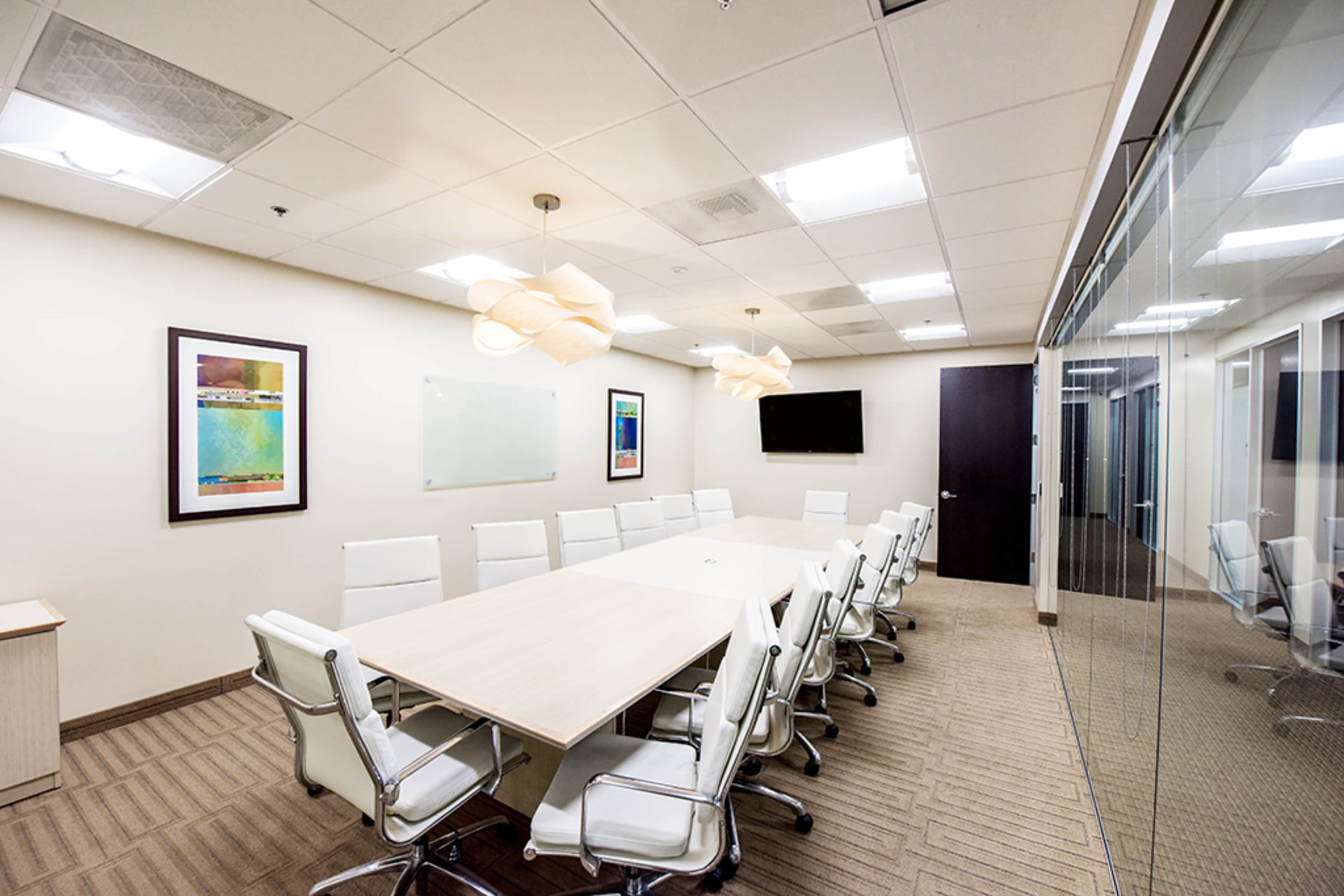 A modern conference room features a long table surrounded by white chairs, with artwork on the walls and large windows separating it from adjoining offices.