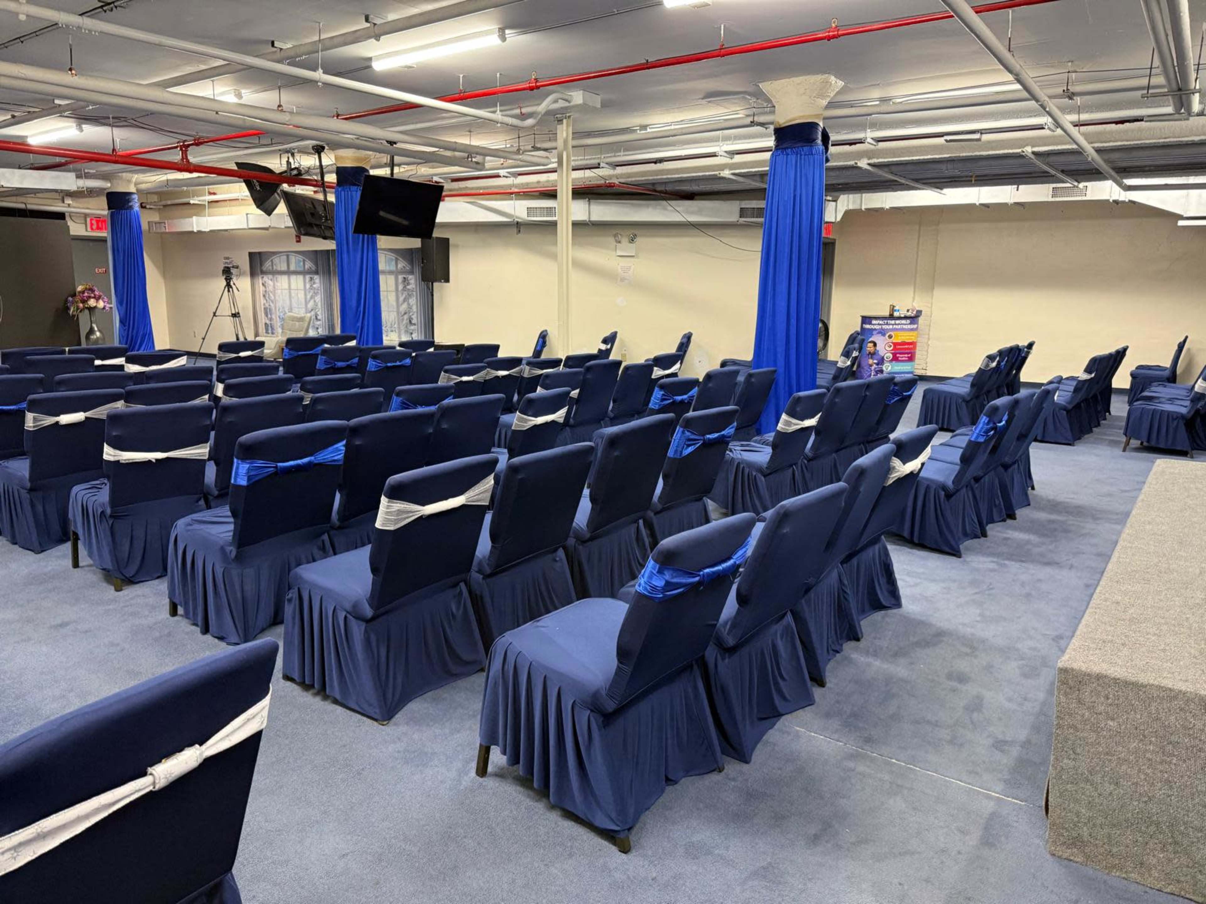 The image shows a spacious room with rows of neatly arranged blue-covered chairs, each adorned with a white sash, and a small stage at the front.