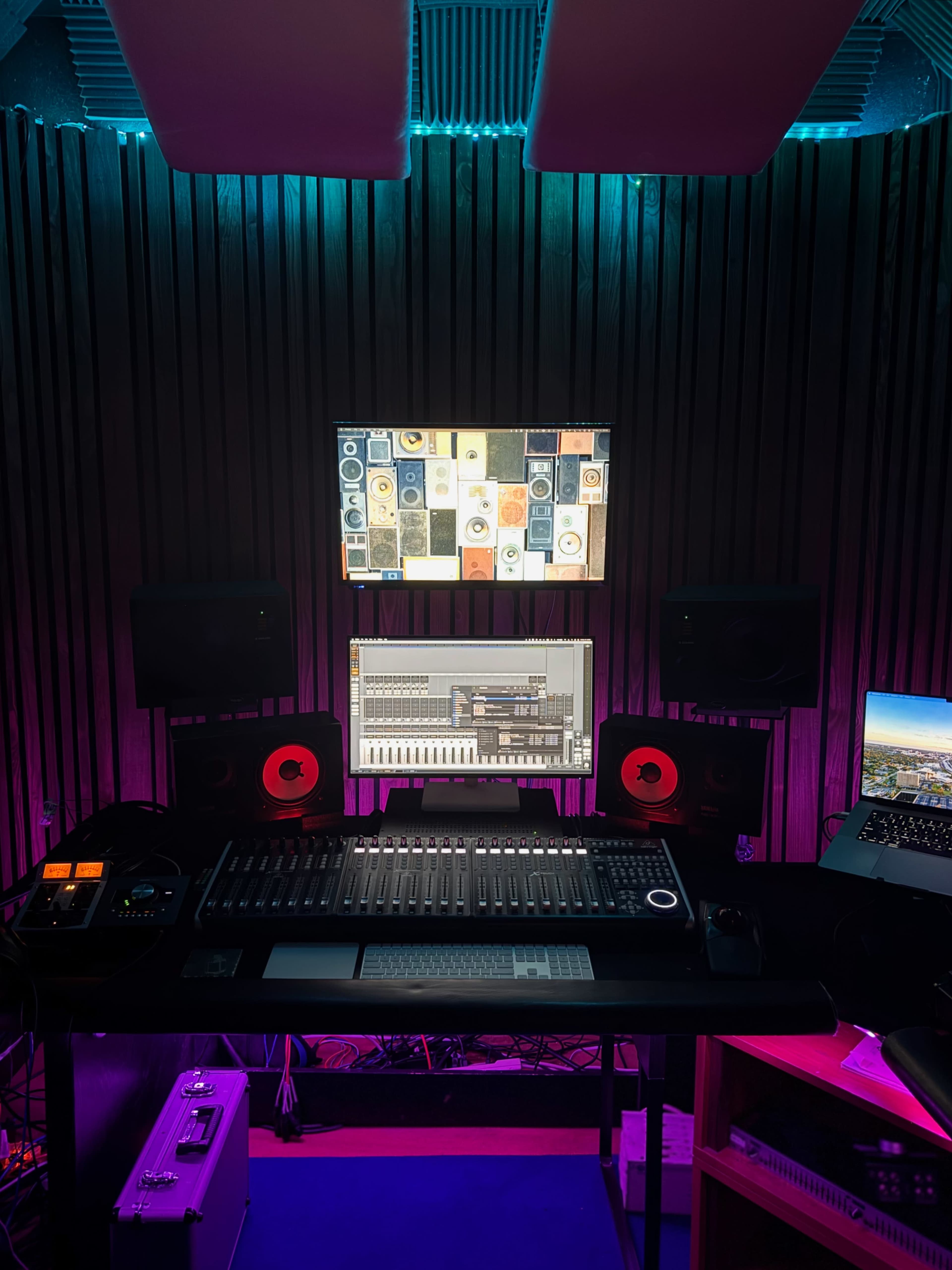 A music studio is set up with a large mixing console, multiple monitors displaying audio software and effects, and speakers placed on either side, illuminated by colorful LED lights.
