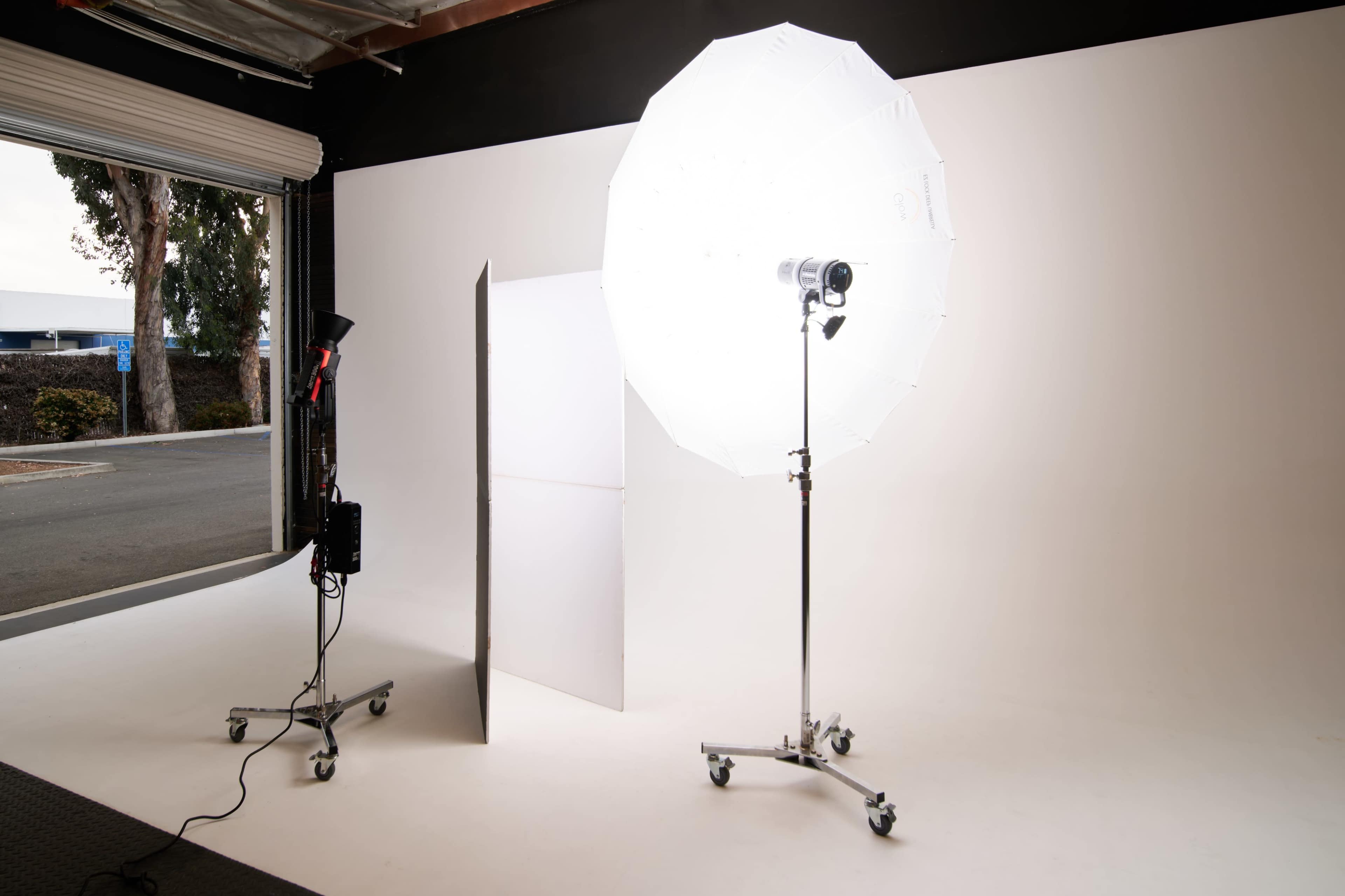 A photography studio setup features a large umbrella light and a reflector stand on a seamless white backdrop.