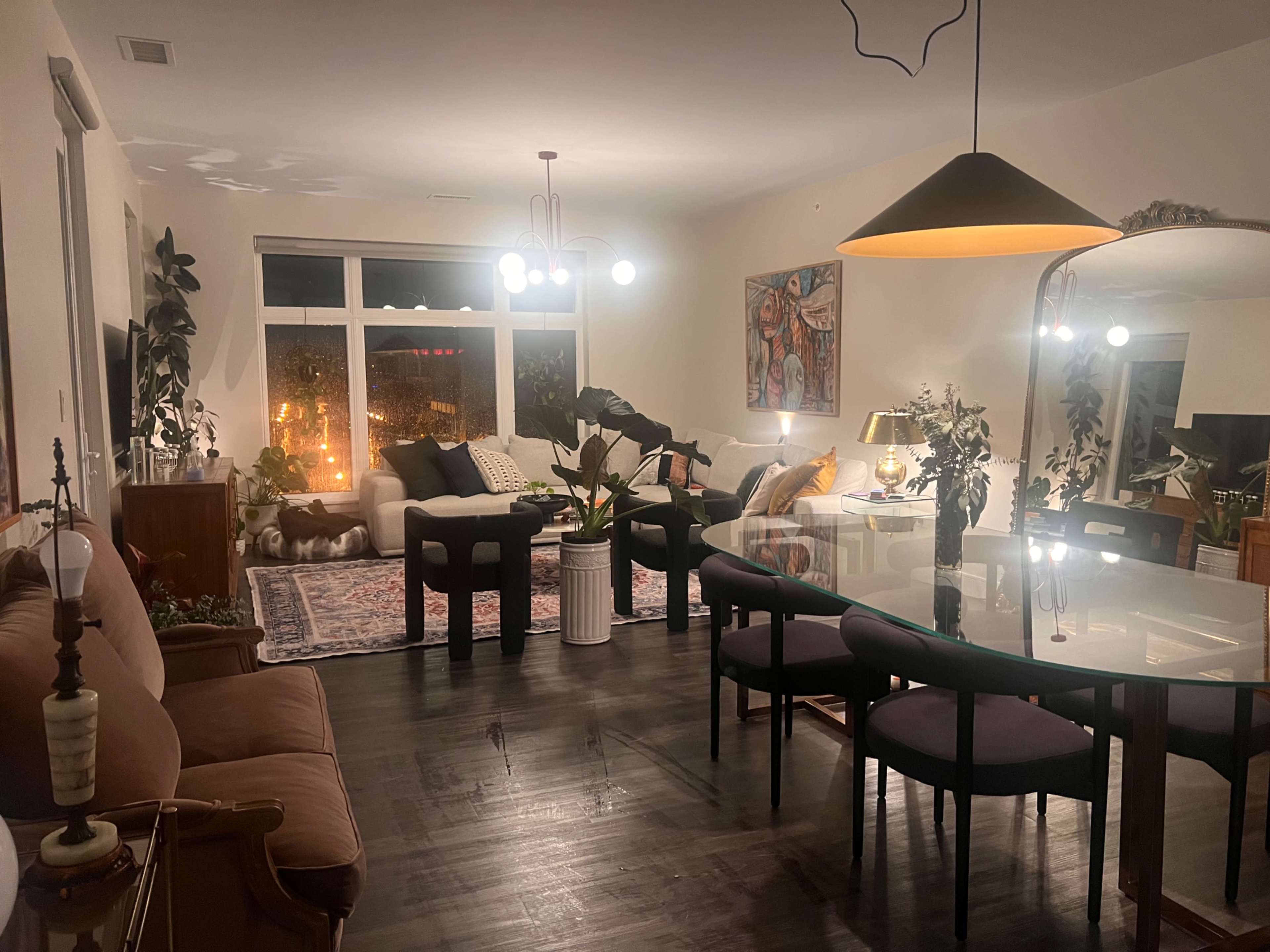 The scene depicts a modern living room with a dining area, featuring a mix of seating arrangements, plants, and decorative artwork.