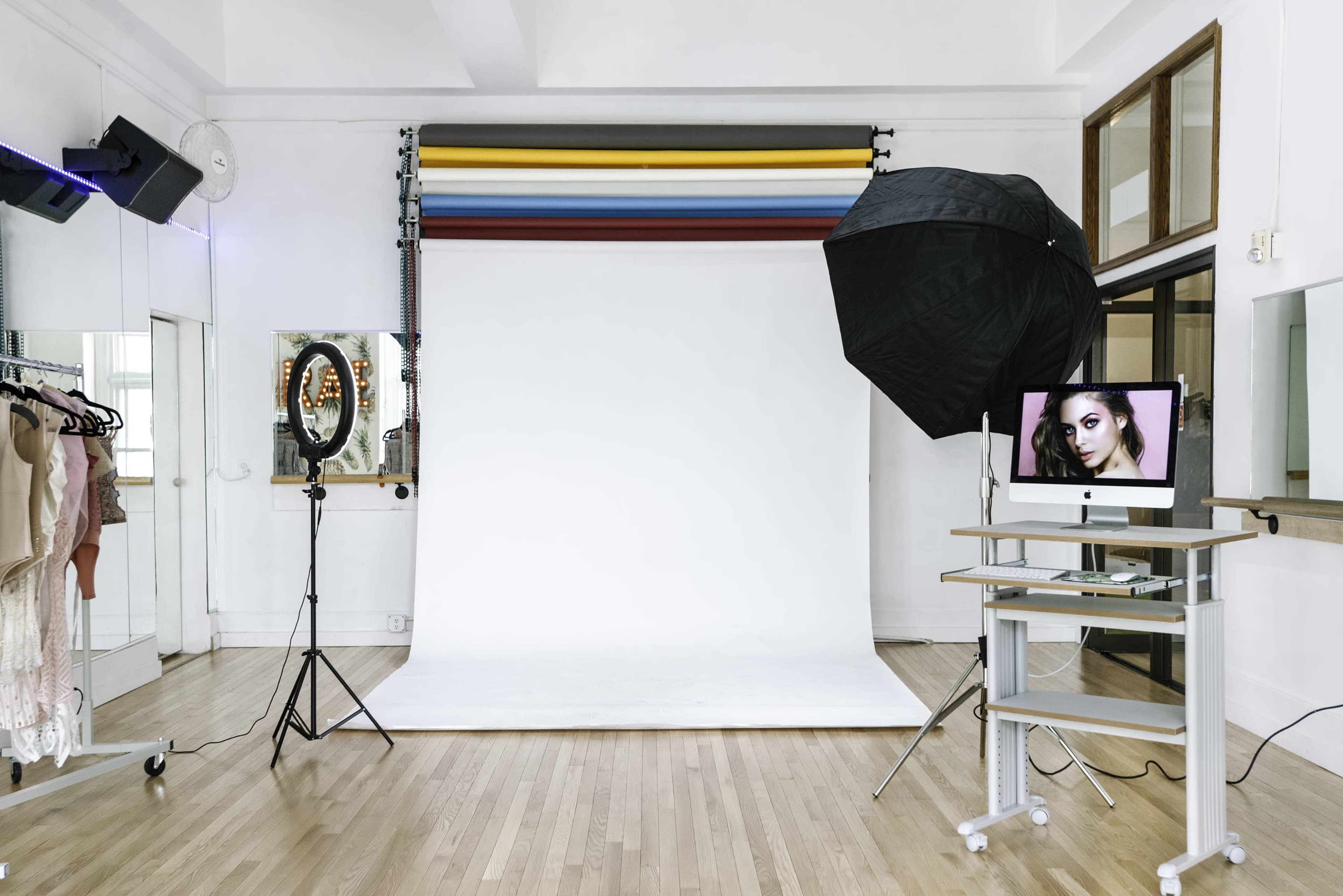 A photography studio with a white backdrop, lighting equipment, a circular mirror, and a computer displaying a model's photograph.