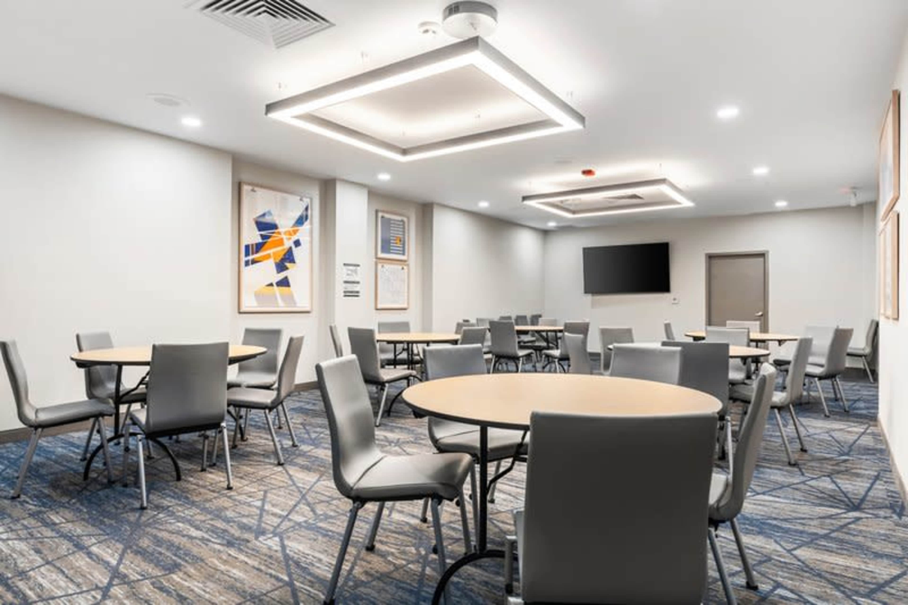The image shows a modern meeting room with several round tables and chairs arranged on a patterned carpet, featuring a television mounted on the wall and artwork.