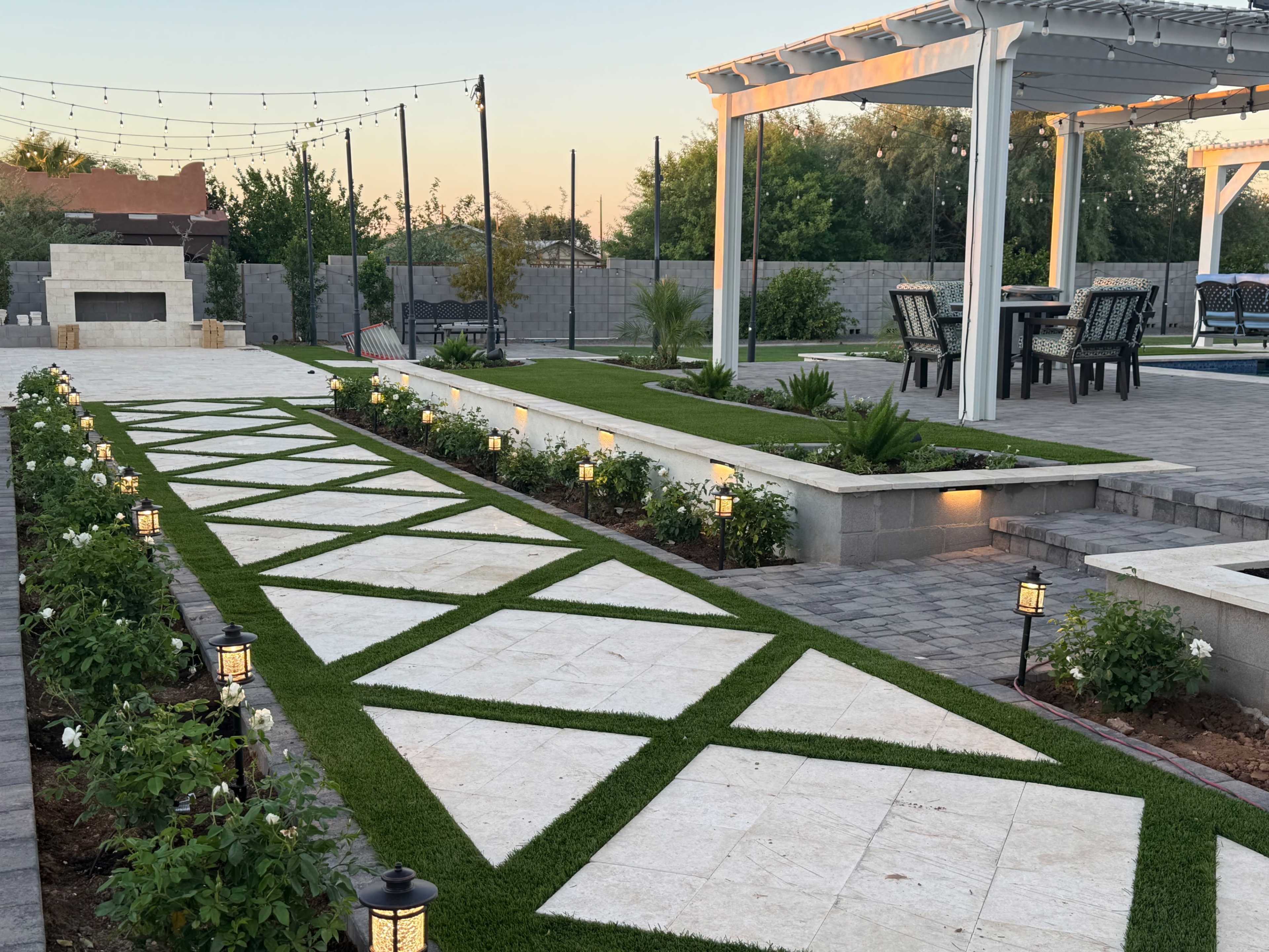 The image shows a landscaped backyard featuring a stone pathway with grass inlay, planters with roses, and a patio area with outdoor furniture and a fireplace.