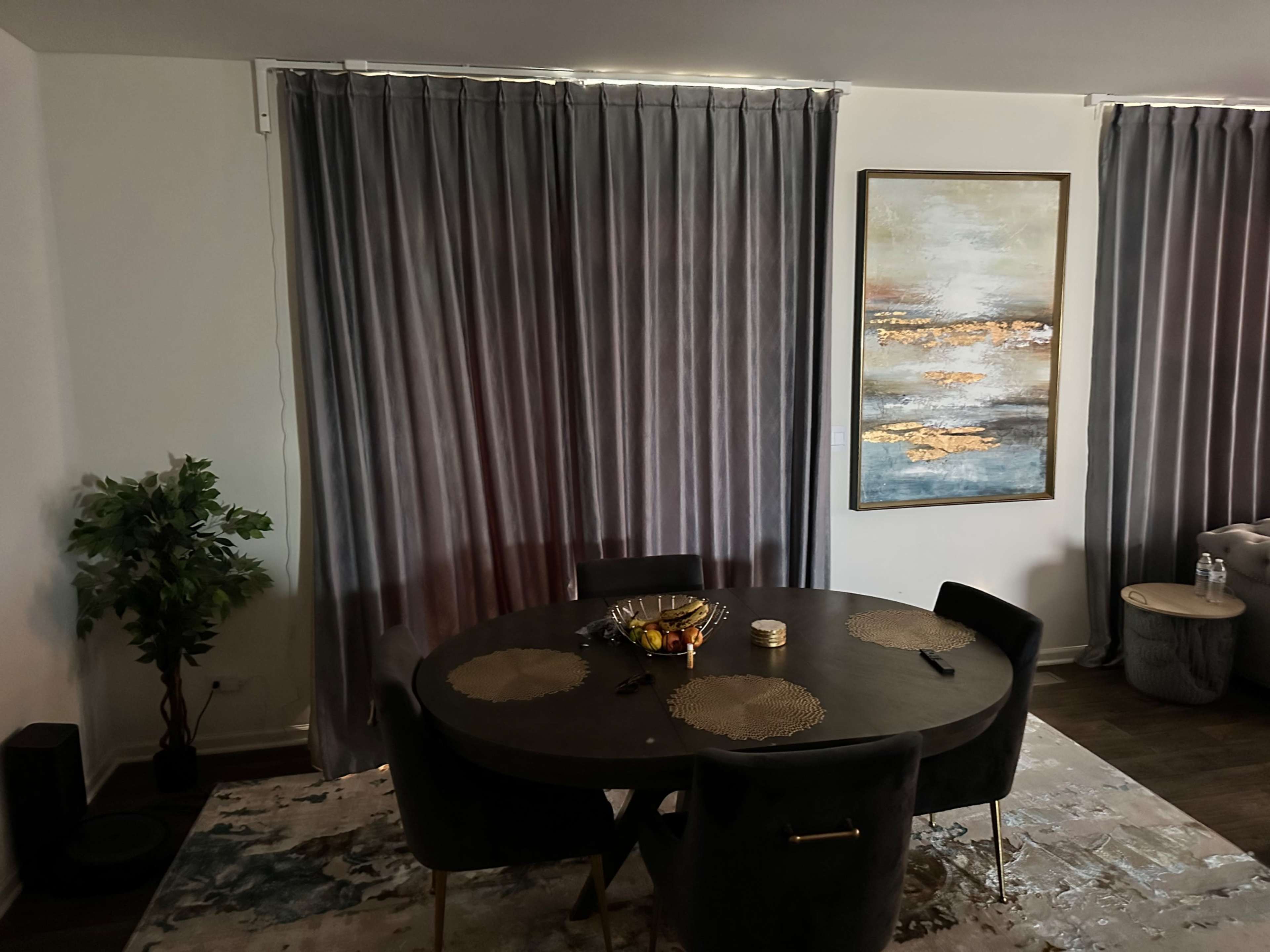 A dining area with a round dark table, four upholstered chairs, a decorative bowl on the table, and curtains framing a large wall art piece beside a potted plant.