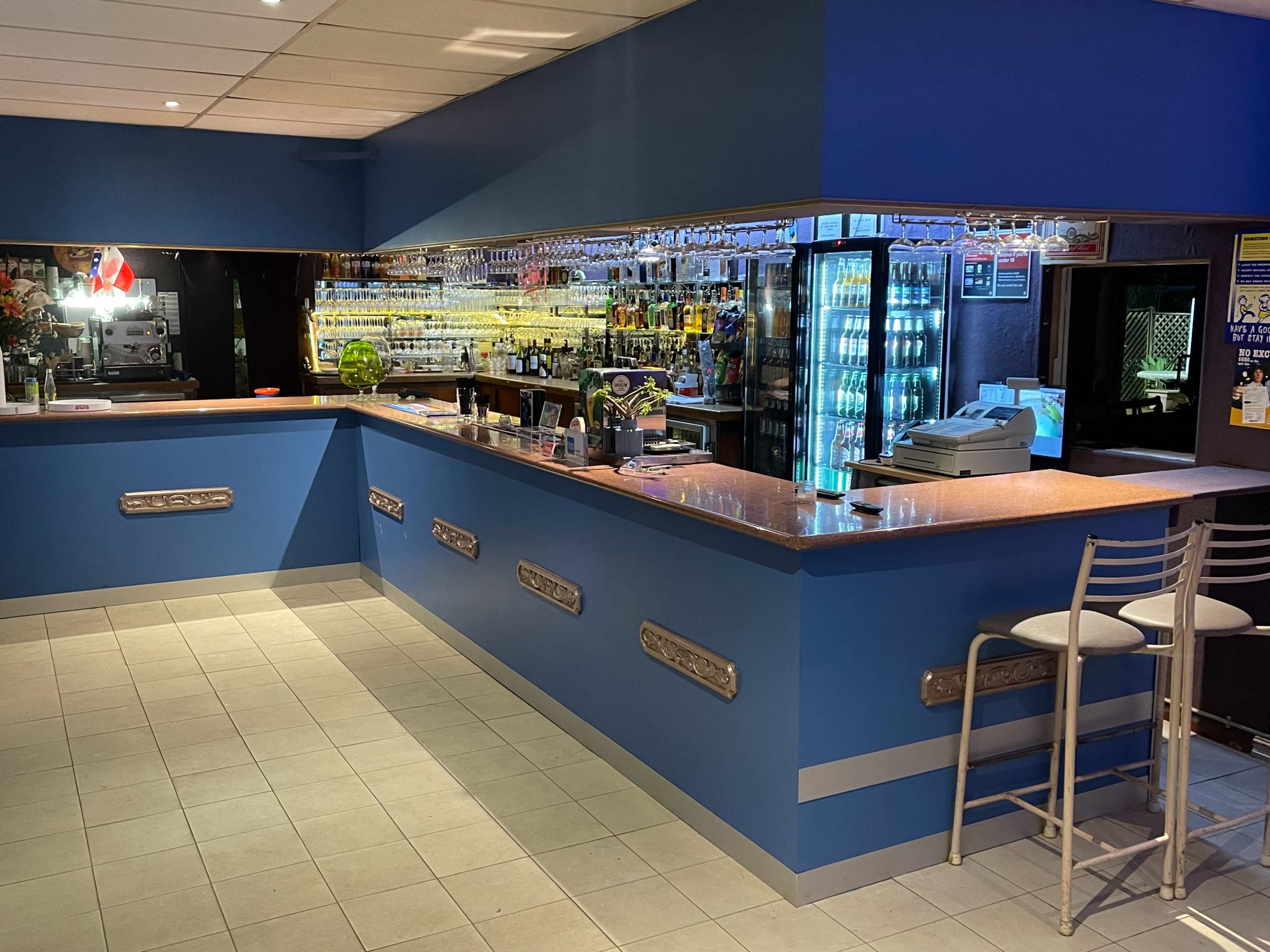 The image shows a brightly lit bar area with a blue wall, a long countertop, and shelves filled with various bottles.