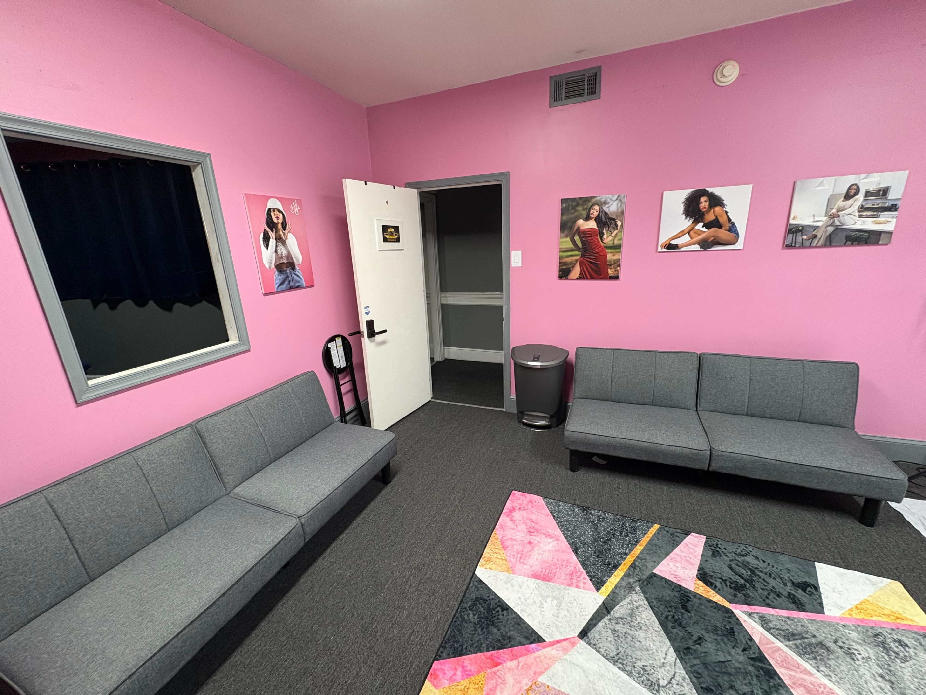 The image shows a brightly colored pink room furnished with gray couches, a rug with geometric patterns, and framed photographs on the walls.