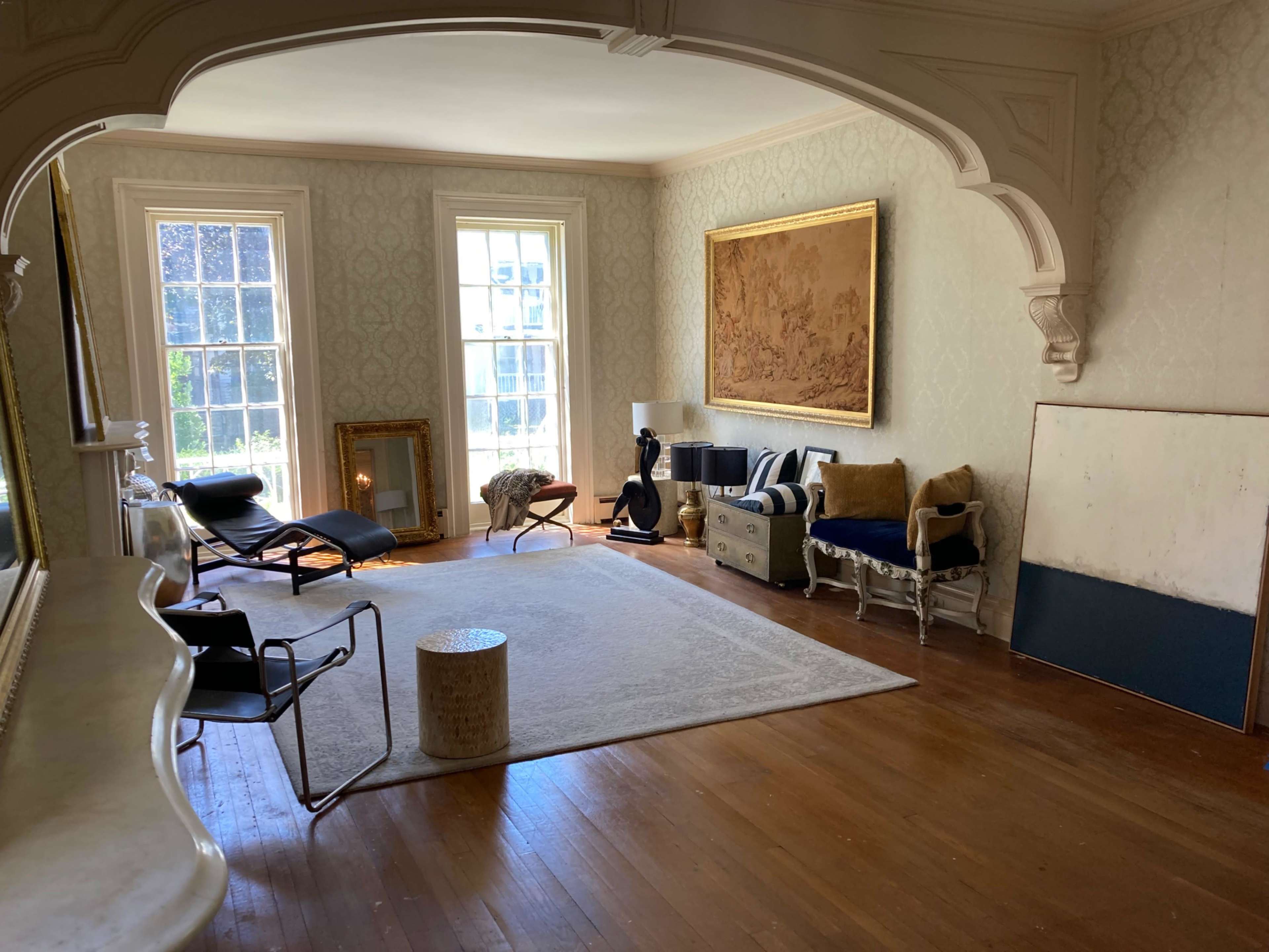 The room features hardwood flooring, large windows, and a variety of furniture including a chair, a lamp, and a large painting on the wall.