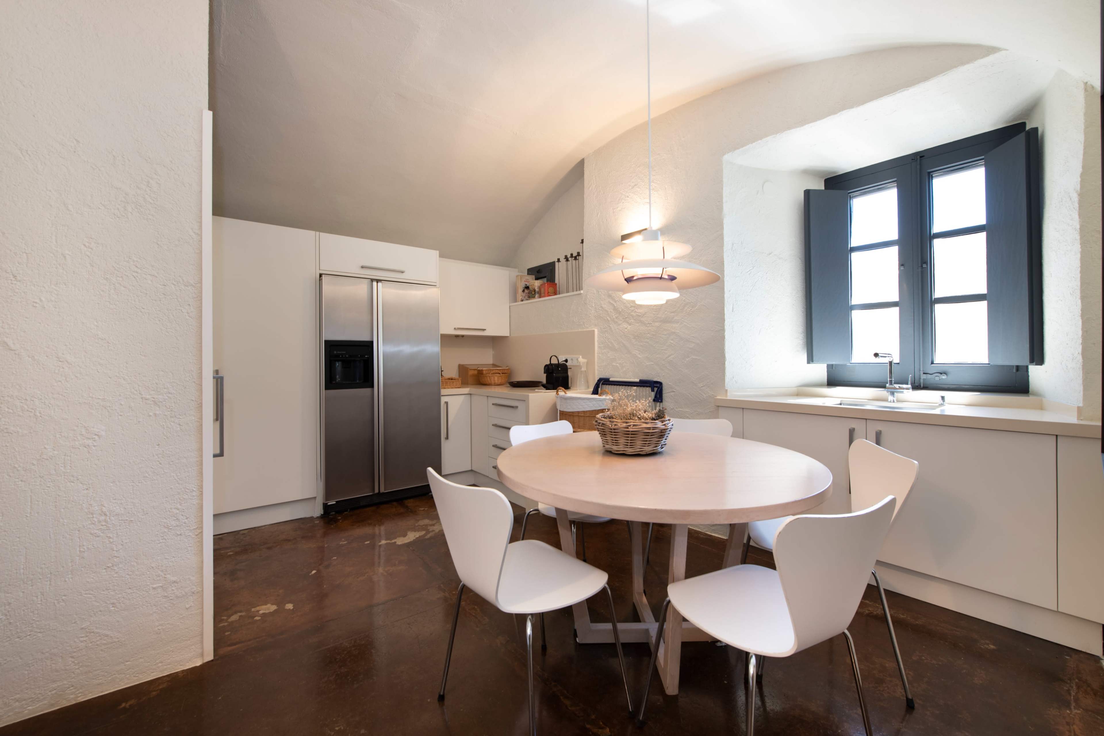 A modern kitchen features a round dining table surrounded by white chairs, with a sleek refrigerator and cabinets visible in the background.