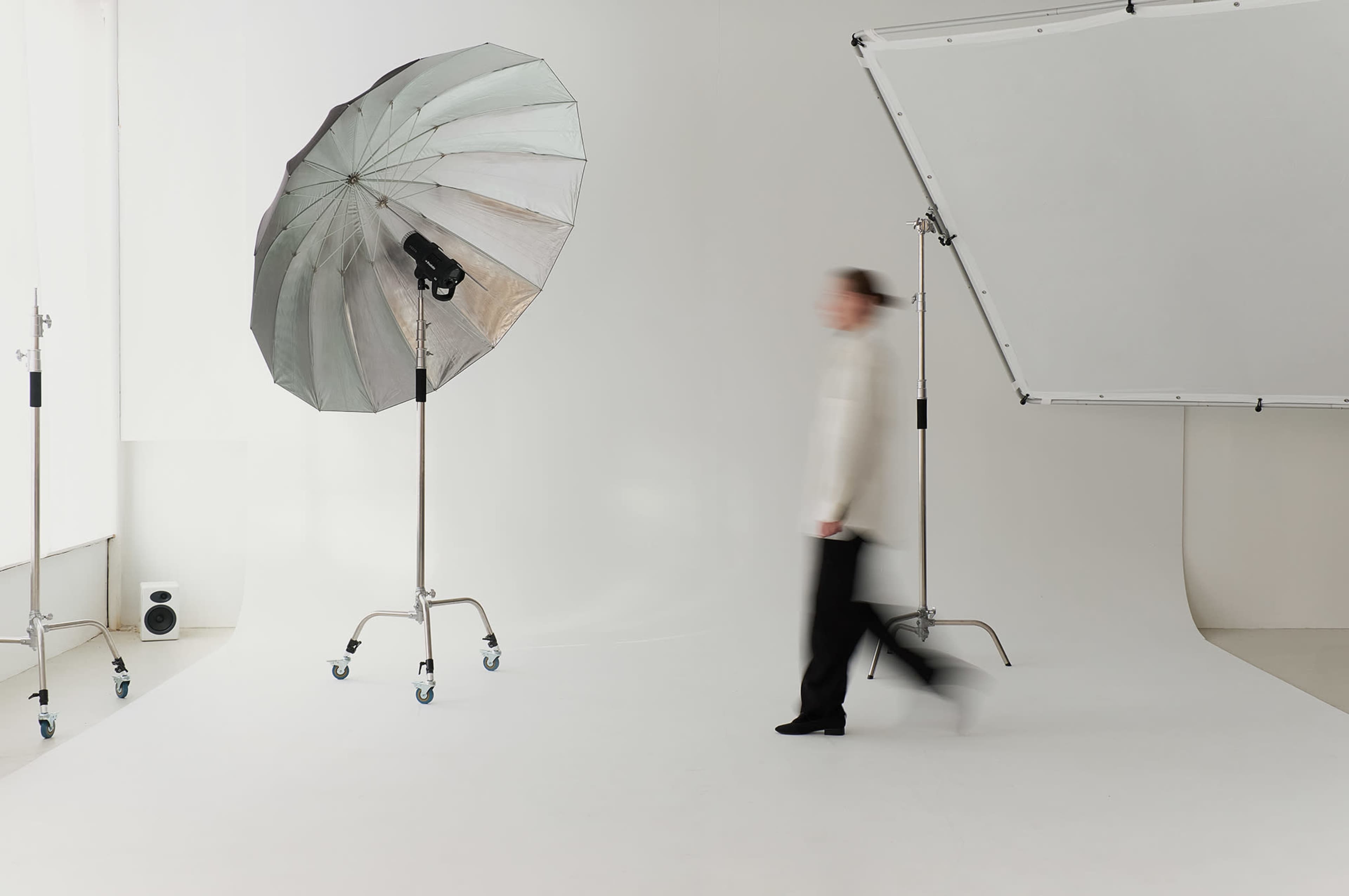 A person walks past a large silver photography umbrella and a backdrop in a bright, minimalistic studio space.