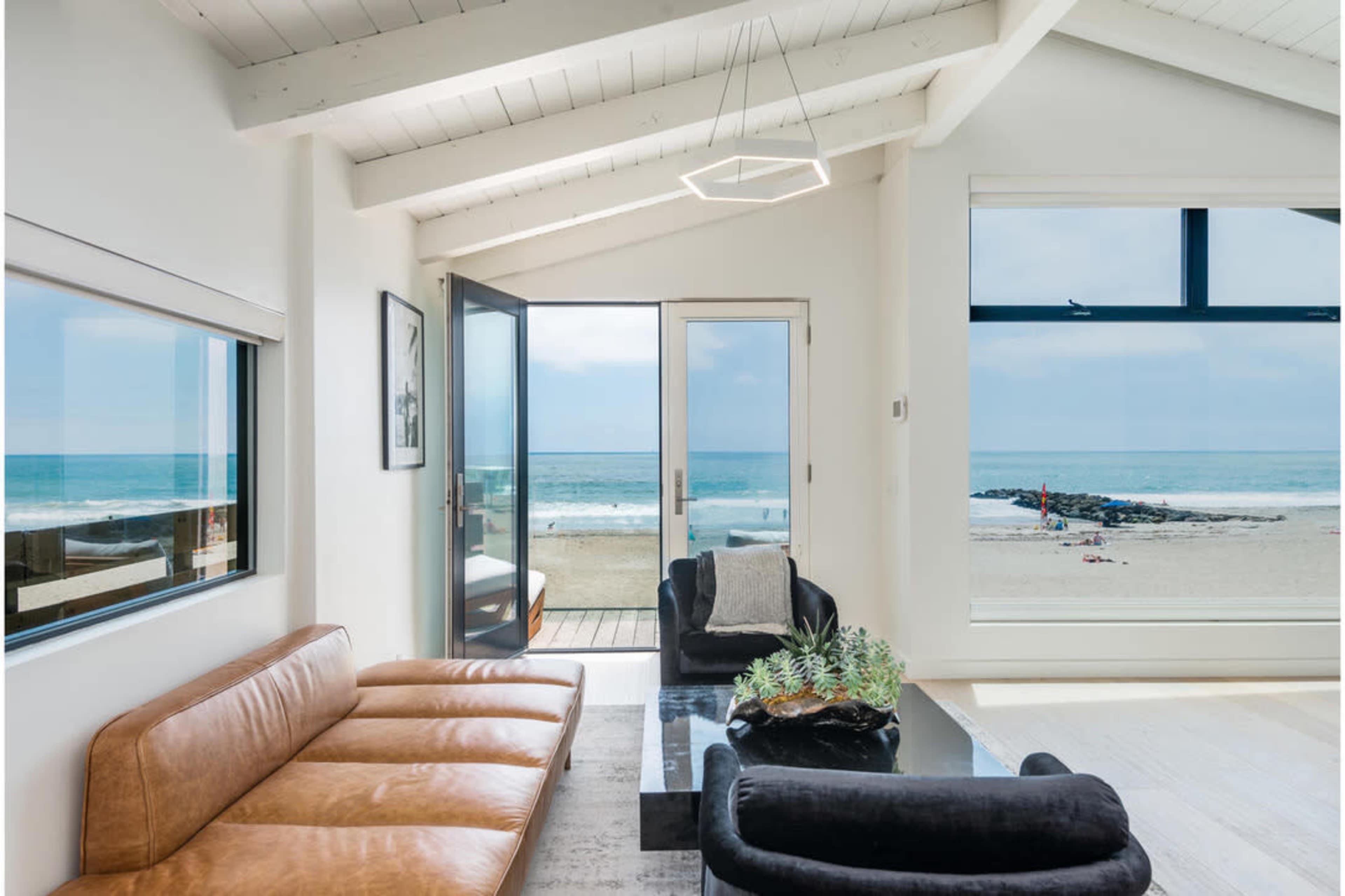 A modern living room features a large window and an open door that overlook a beach with waves and people in the distance.
