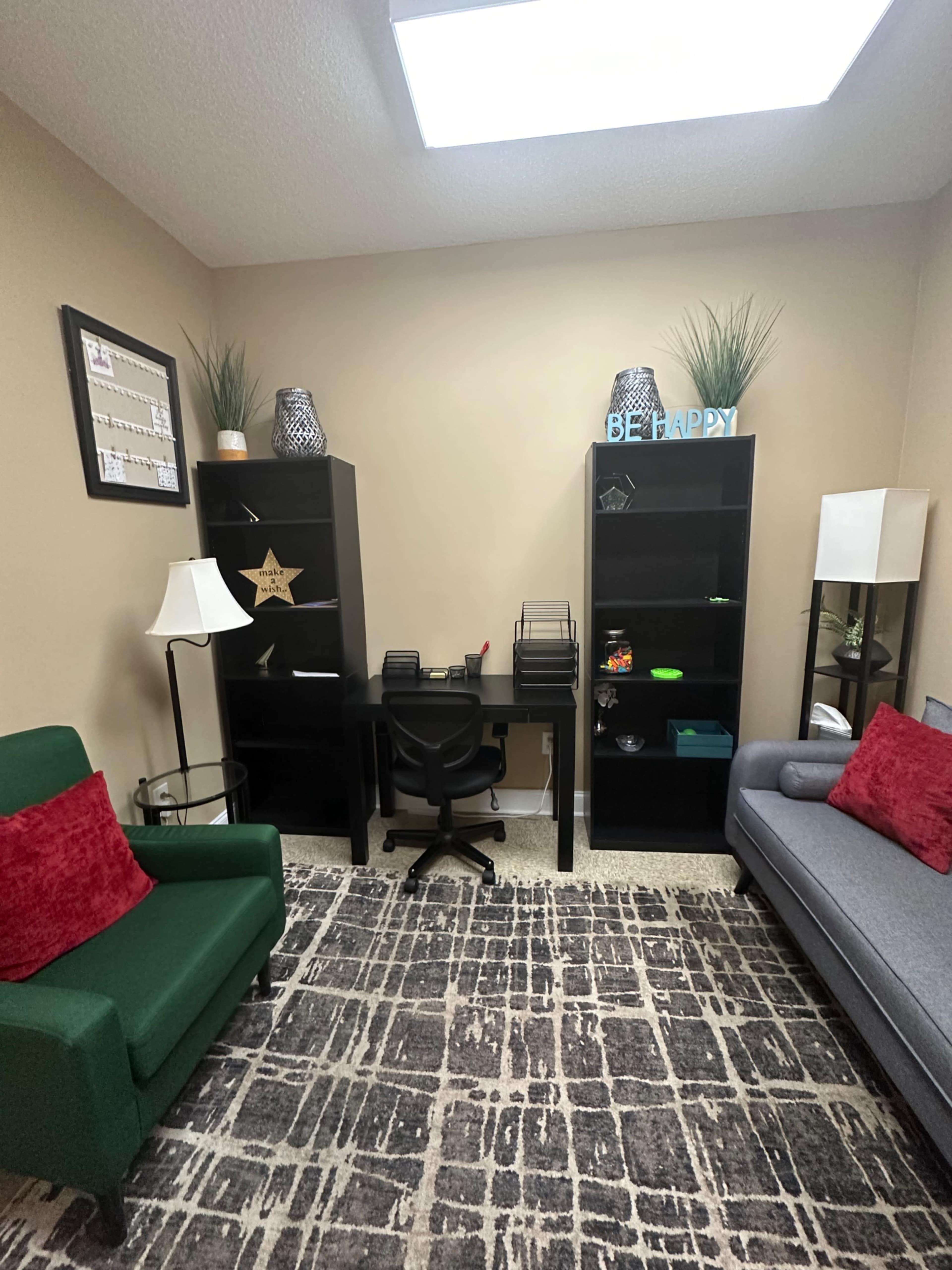 The image shows a small office space with two bookshelves, a desk with a chair, and a couch and armchair arranged around a patterned rug.
