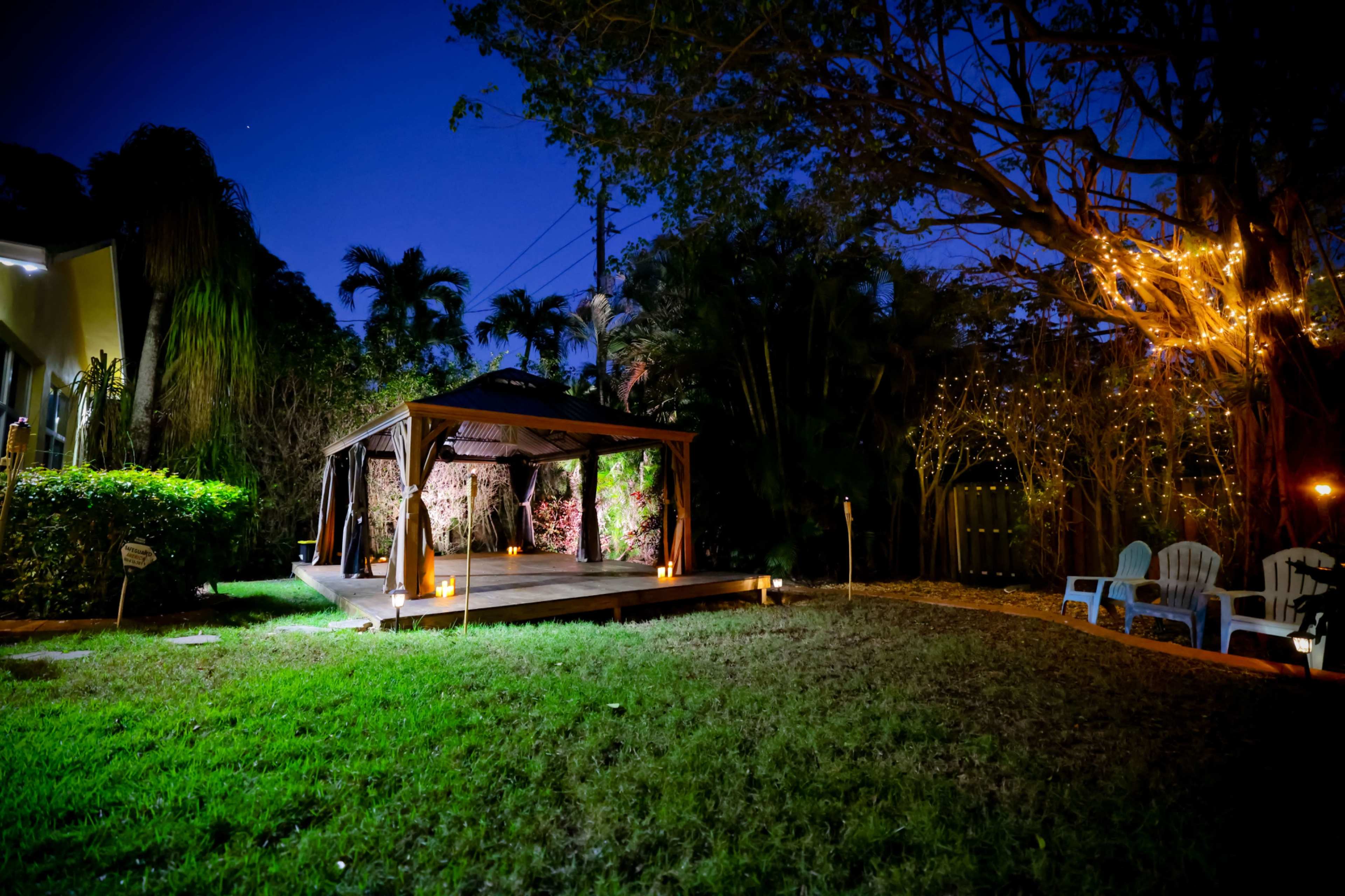 A wooden gazebo is illuminated by lanterns in a grassy yard surrounded by trees and decorated with string lights.