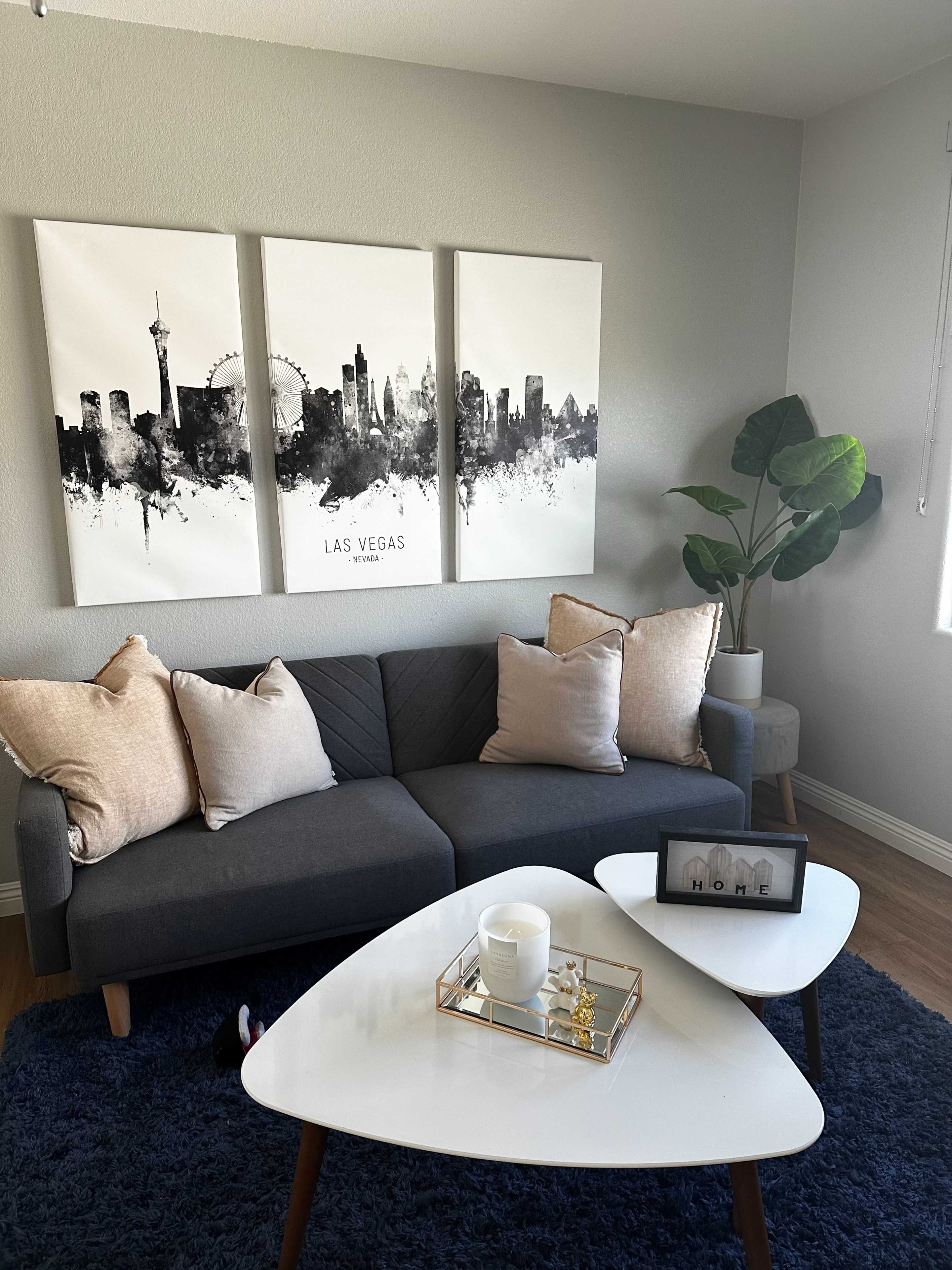 The image shows a modern living room with a gray sofa adorned with decorative pillows, a coffee table, and three framed cityscape prints featuring Las Vegas on the wall.