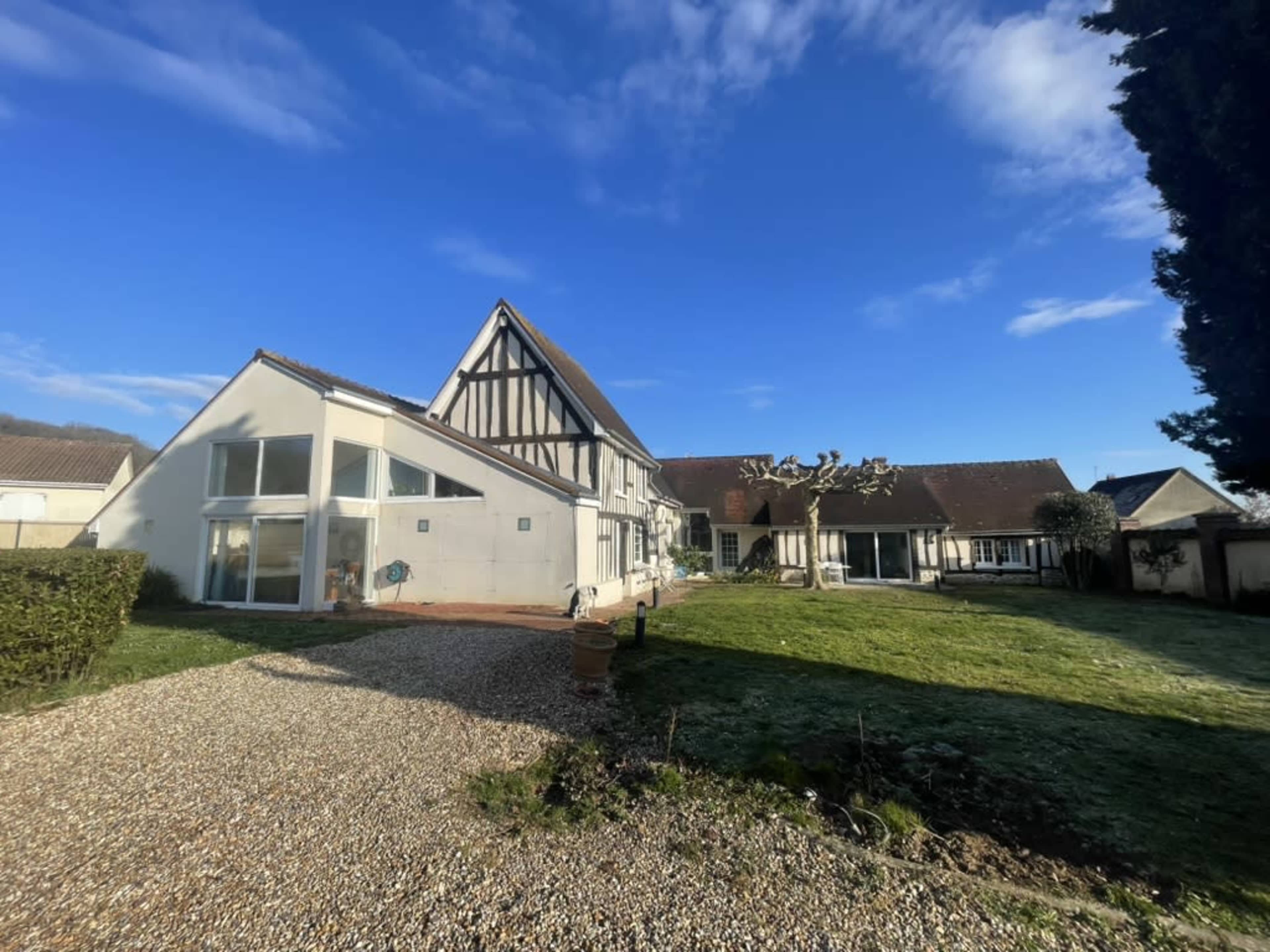 The image shows a large house with a mix of modern and traditional architectural features, set in a grassy area under a clear blue sky.