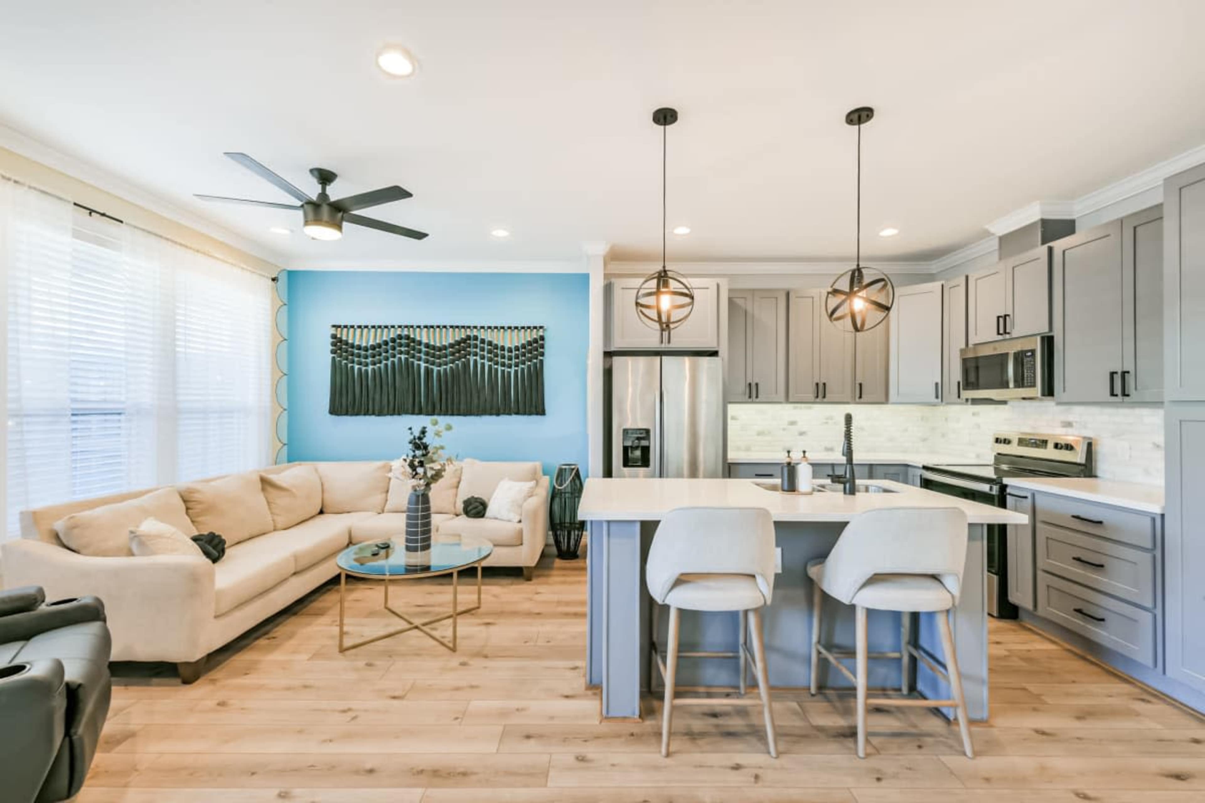 The image shows a modern kitchen and living area featuring a beige sectional sofa, a glass coffee table, and a kitchen island with bar stools, all set against light-colored cabinetry and a blue accent wall.