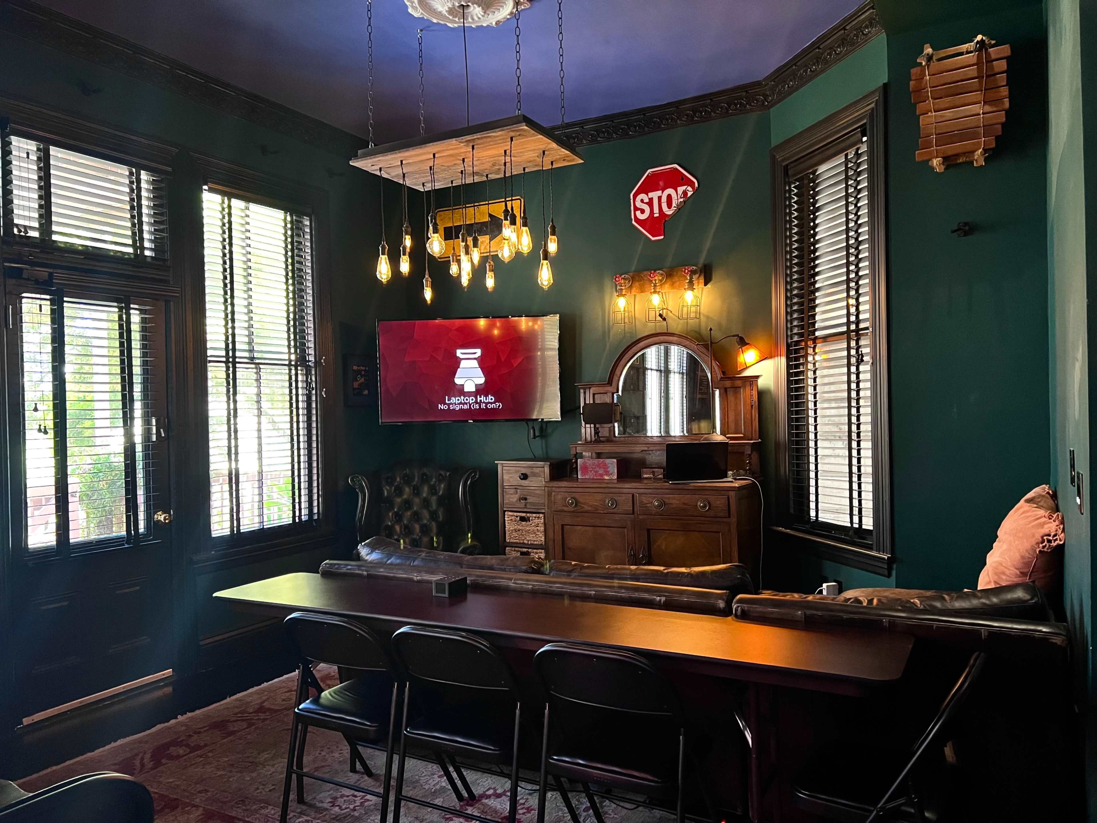 A cozy room with dark green walls, vintage furniture, a television mounted on the wall, and a long table with folding chairs arranged in front.