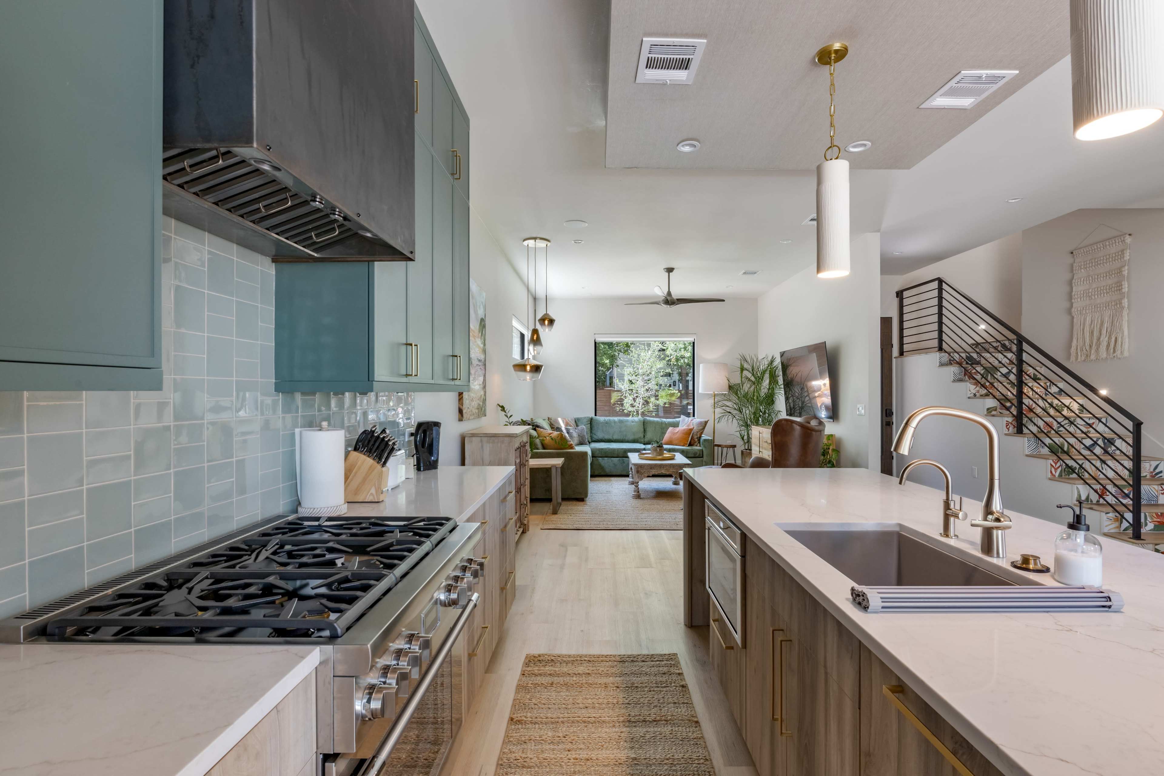 The image shows a modern kitchen with a gas stove, green and wooden cabinetry, an island with a sink, and a view of a living area with a sofa and plants.