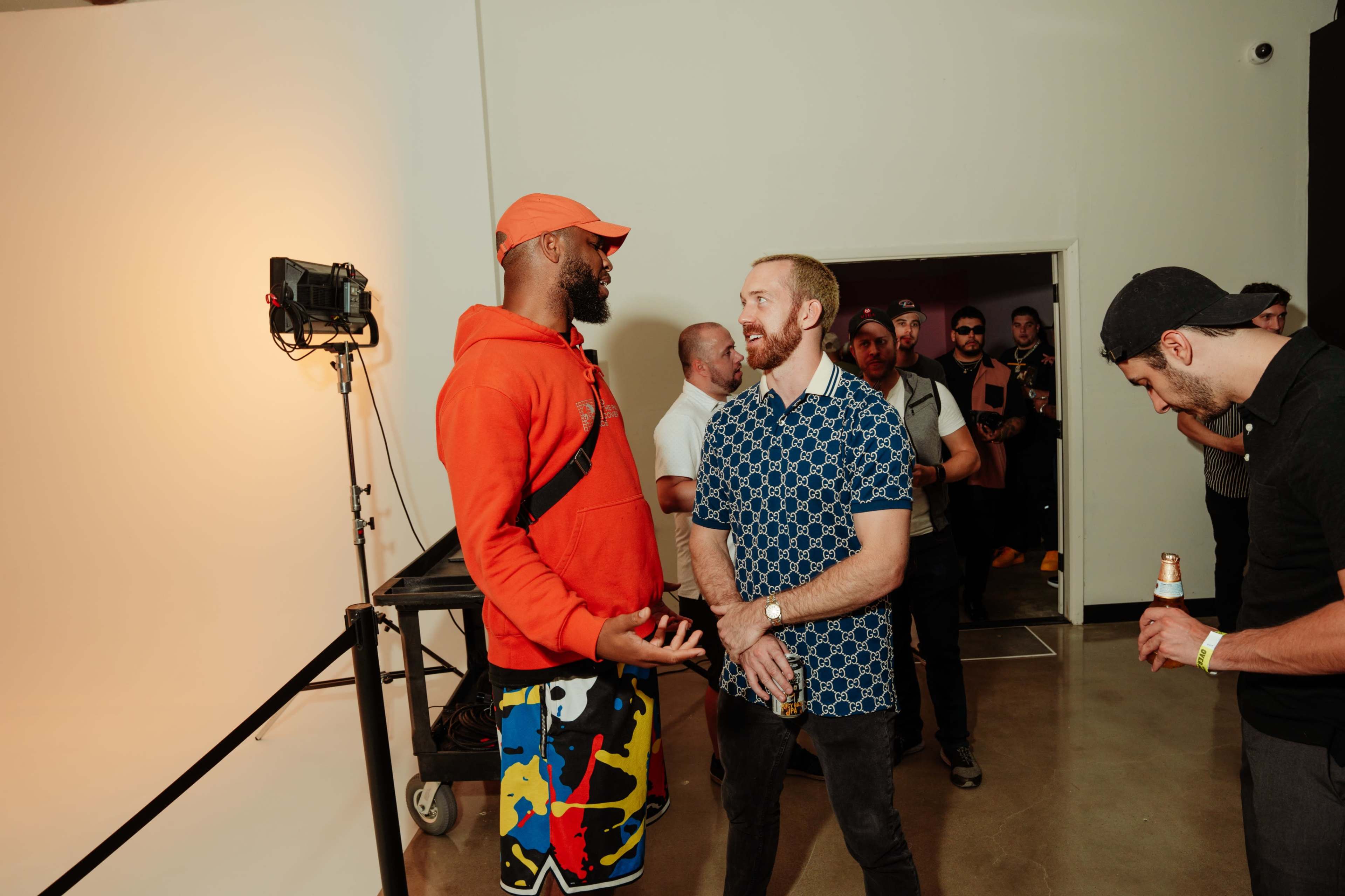 Two men engage in conversation in front of a group of people waiting in line at an event, with a large light setup in the background.