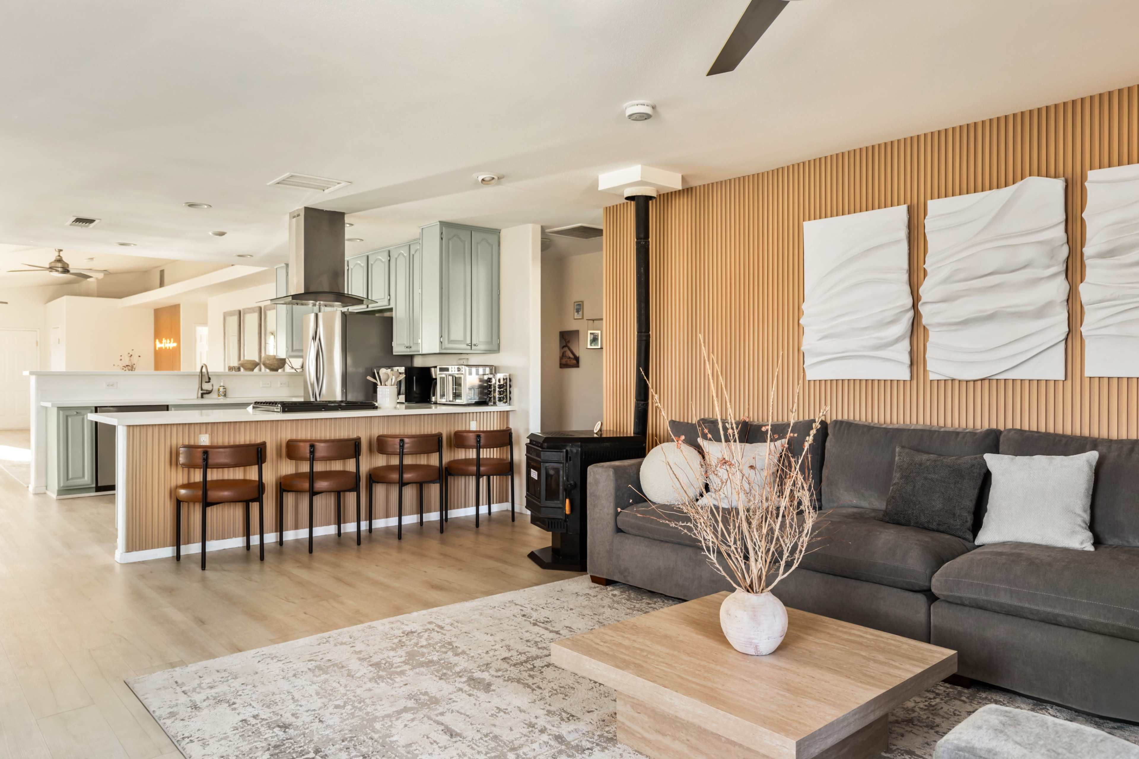 The image showcases a modern interior space featuring a living area with a gray sofa, a wooden coffee table, and a kitchen bar with four high stools.