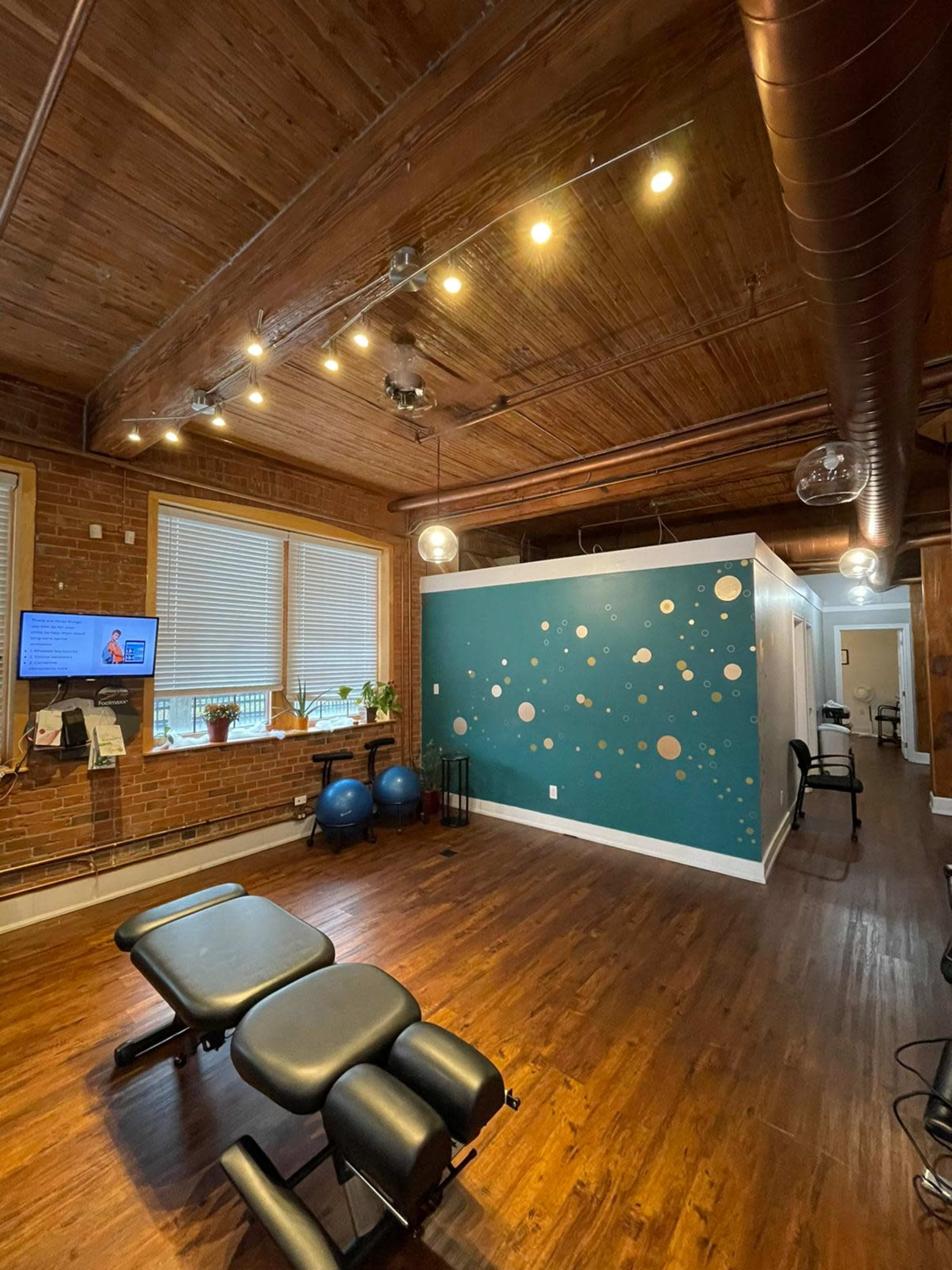 A well-lit therapy room with wooden floors, a dark green accent wall featuring circular designs, and several pieces of exercise equipment.