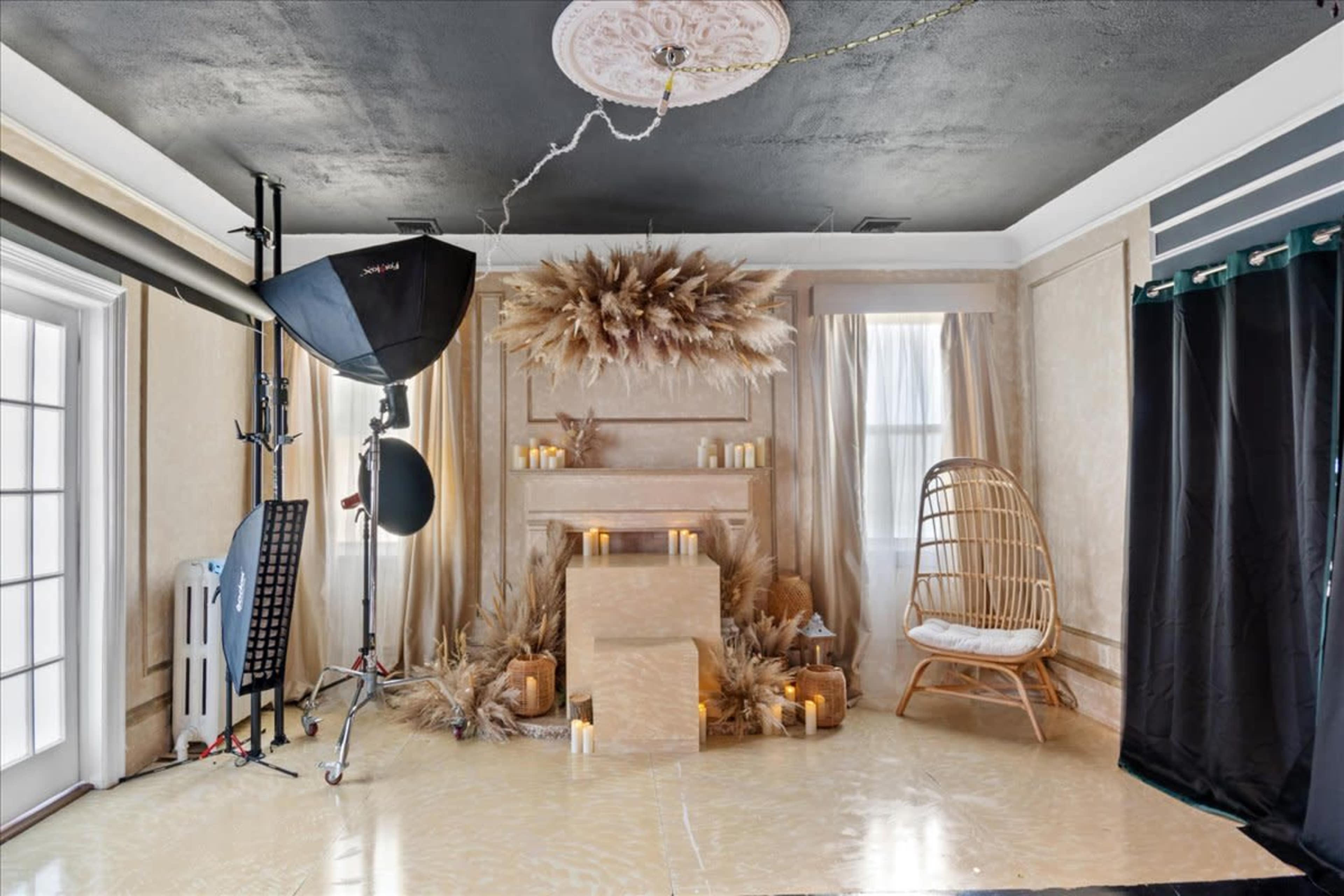 A softly lit studio setup featuring a mantle adorned with candles and decorative elements, a rattan chair, and a large light source on a stand.