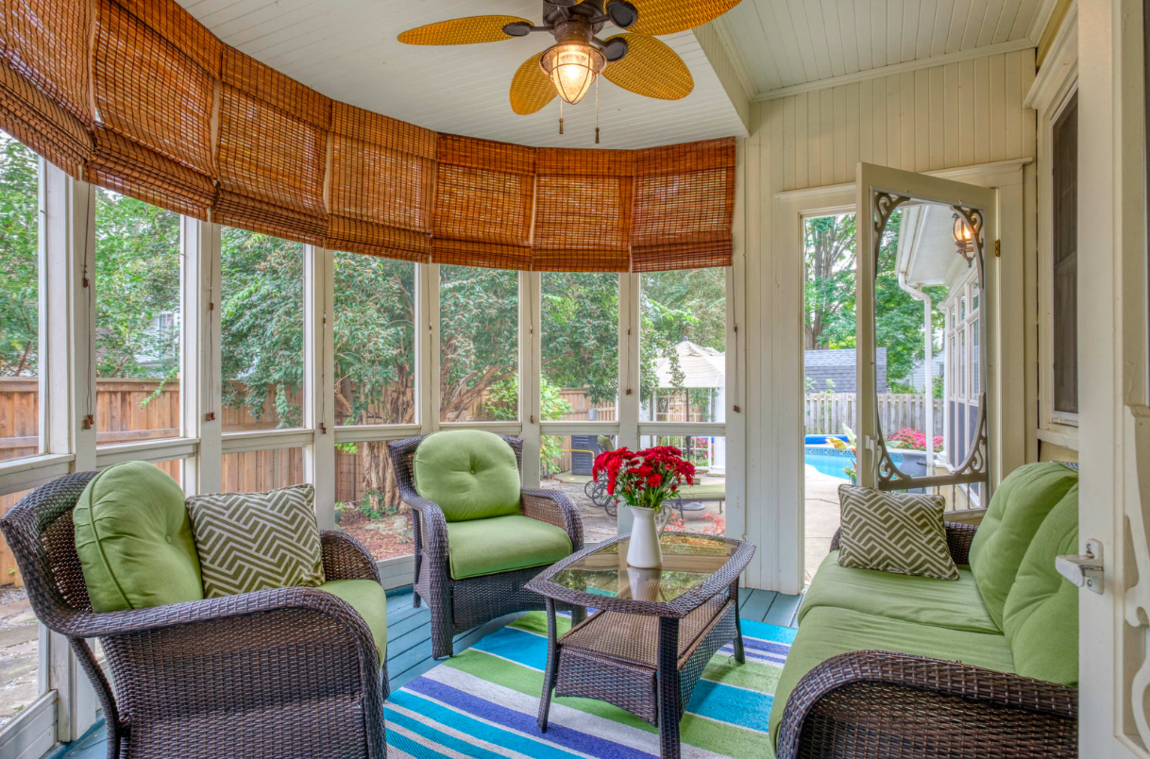 A sunlit screened-in porch furnished with wicker chairs, a small table, a ceiling fan, and a vibrant rug.